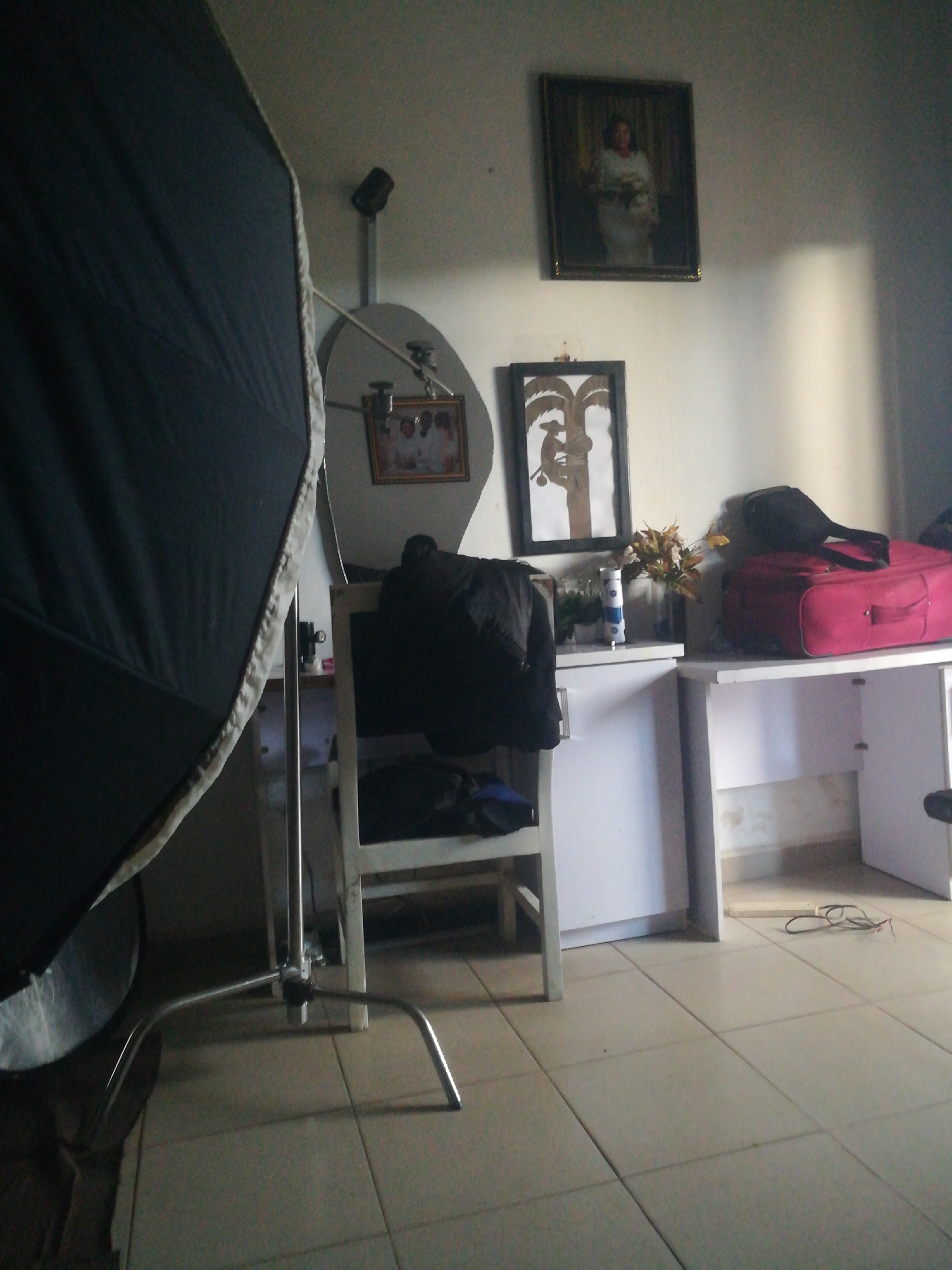 The image depicts an indoor scene with a large black photography umbrella dominating the left side of the frame. The background reveals a room with tiled flooring and light-colored walls. A white desk is positioned against the wall, with a pink suitcase and a black bag resting on its surface. To the left of the desk is a white cabinet with items on top, including a vase with artificial flowers and a spray can. Two framed pictures hang on the wall above the desk; one is a portrait of a woman and the other contains smaller framed photographs. A white chair with a black garment draped over it stands in front of the cabinet. The overall impression is that of a room that might be used for photography or storage, with a somewhat cluttered but functional arrangement of objects.