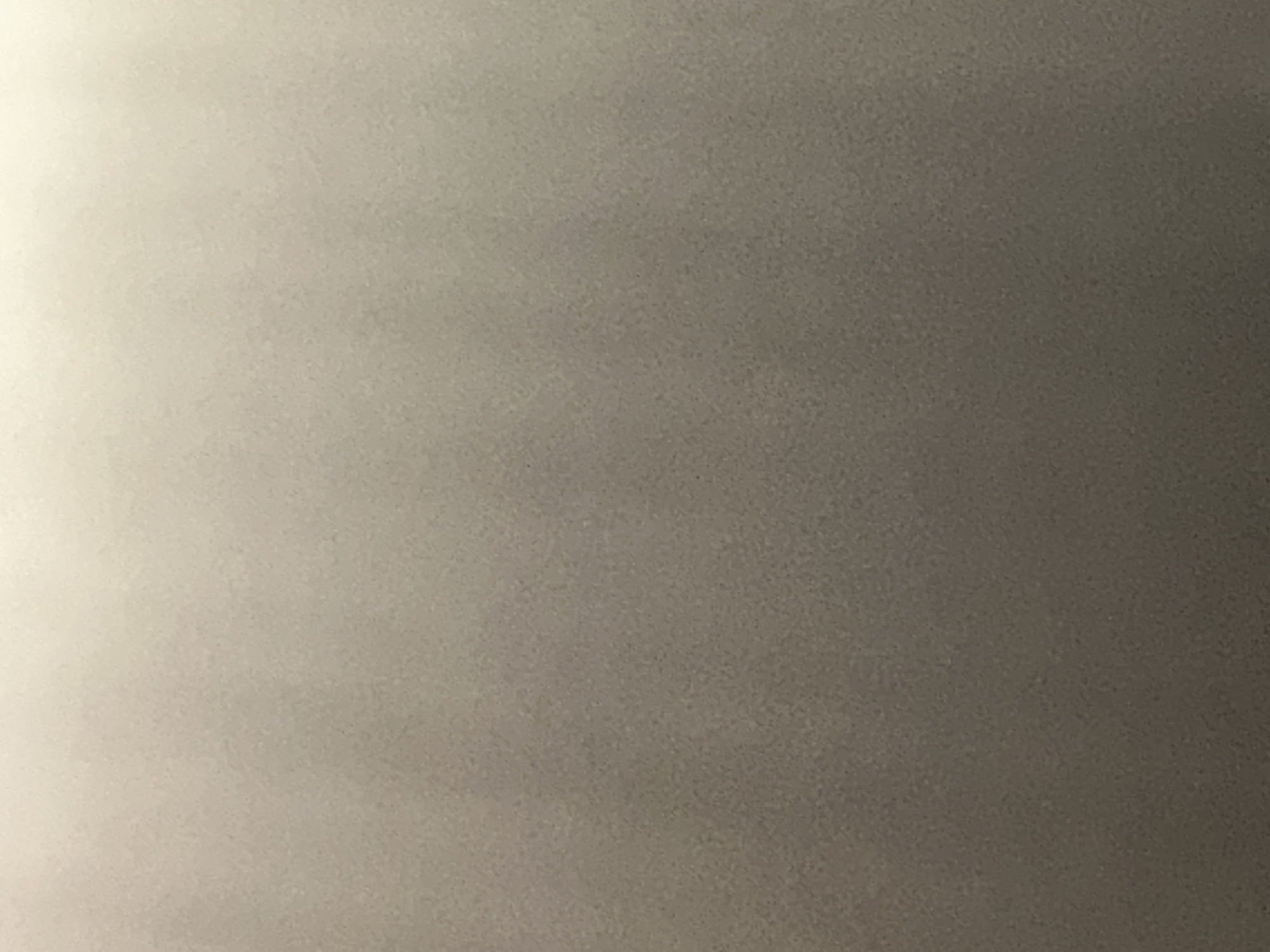 The image is a close-up, abstract shot of a textured surface that appears to be a wall or ceiling. There are no discernible subjects like people or objects, nor is there any clear indication of a scene or setting beyond a generic indoor environment. The lighting in the image is uneven, with a bright, white light source illuminating the top left corner, creating a gradient that fades to a darker, grayish tone towards the bottom right. This suggests the photo might have been taken during the daytime, possibly with artificial lighting contributing to the bright area. The texture of the surface is granular and appears slightly uneven, with subtle variations in shade and color, giving it a matte finish. There is no visible text in the image. The context of Belgrade, Serbia, does not provide any specific visual cues within the image itself, as the surface is nondescript and could be found anywhere. The overall impression is one of simplicity and a lack of detail, focusing solely on the interplay of light and shadow on a textured surface.