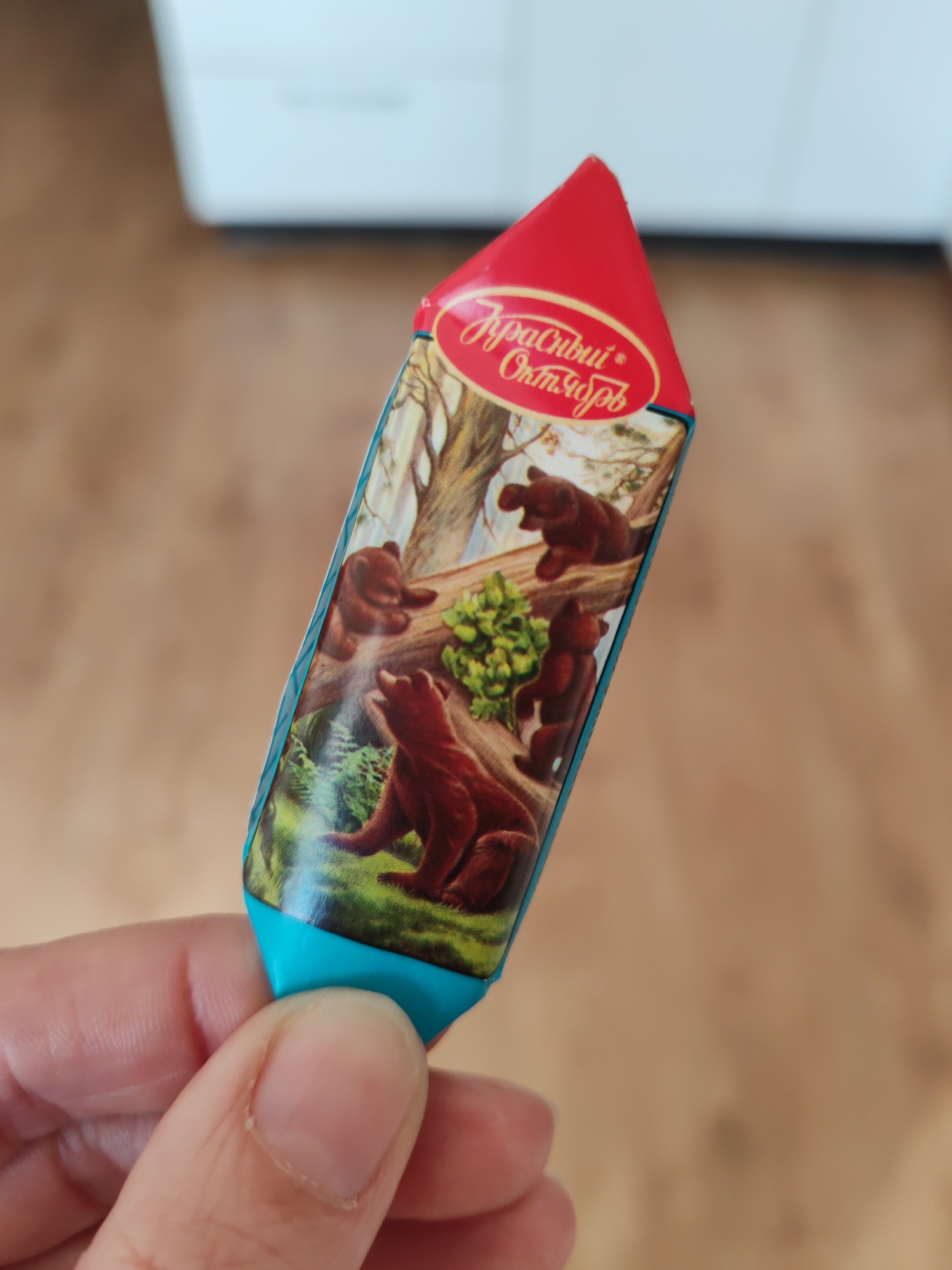 A close-up, eye-level shot features a light-skinned hand holding a distinctively shaped candy or chocolate wrapper. The wrapper has a pointed red top and a blue base, resembling a pencil or a small rocket. The main body of the wrapper displays a classic illustration: several brown bears in a forest setting, with one bear on a fallen log and others on the ground amidst trees and foliage. This is the iconic design for "Mishka Kosolapy" (Clubfoot Bear) chocolate.
Visible text in Cyrillic on the red top of the wrapper reads "Красный Октябрь" (Krasny Oktyabr), which translates to "Red October," identifying a renowned Russian confectionery factory. The hand holds the wrapper gently between the thumb and index finger. The background is softly blurred, suggesting an indoor environment with a light-colored surface in the upper left and a warm, medium-brown surface, possibly a wooden floor or table, to the right. The scene captures a simple, everyday moment, characteristic of life in Saint Petersburg, Russia, given the nature of the confectionery.