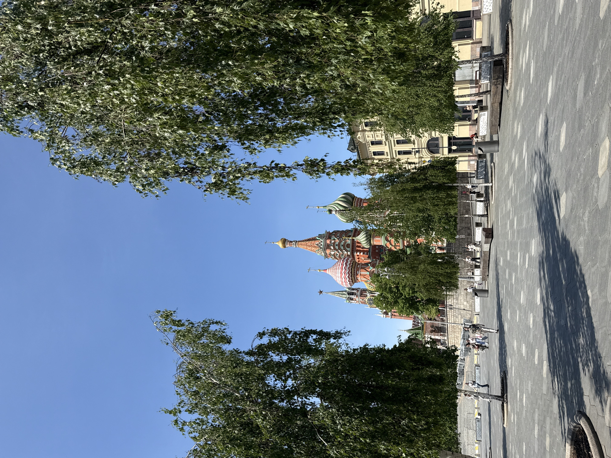 The image depicts St. Basil's Cathedral in Moscow, Russia, partially obscured by trees. The sky is a clear blue. A paved area, likely Red Square, is visible with street furniture and a few pedestrians. Shadows indicate a sunny day. The image is taken from a low angle.