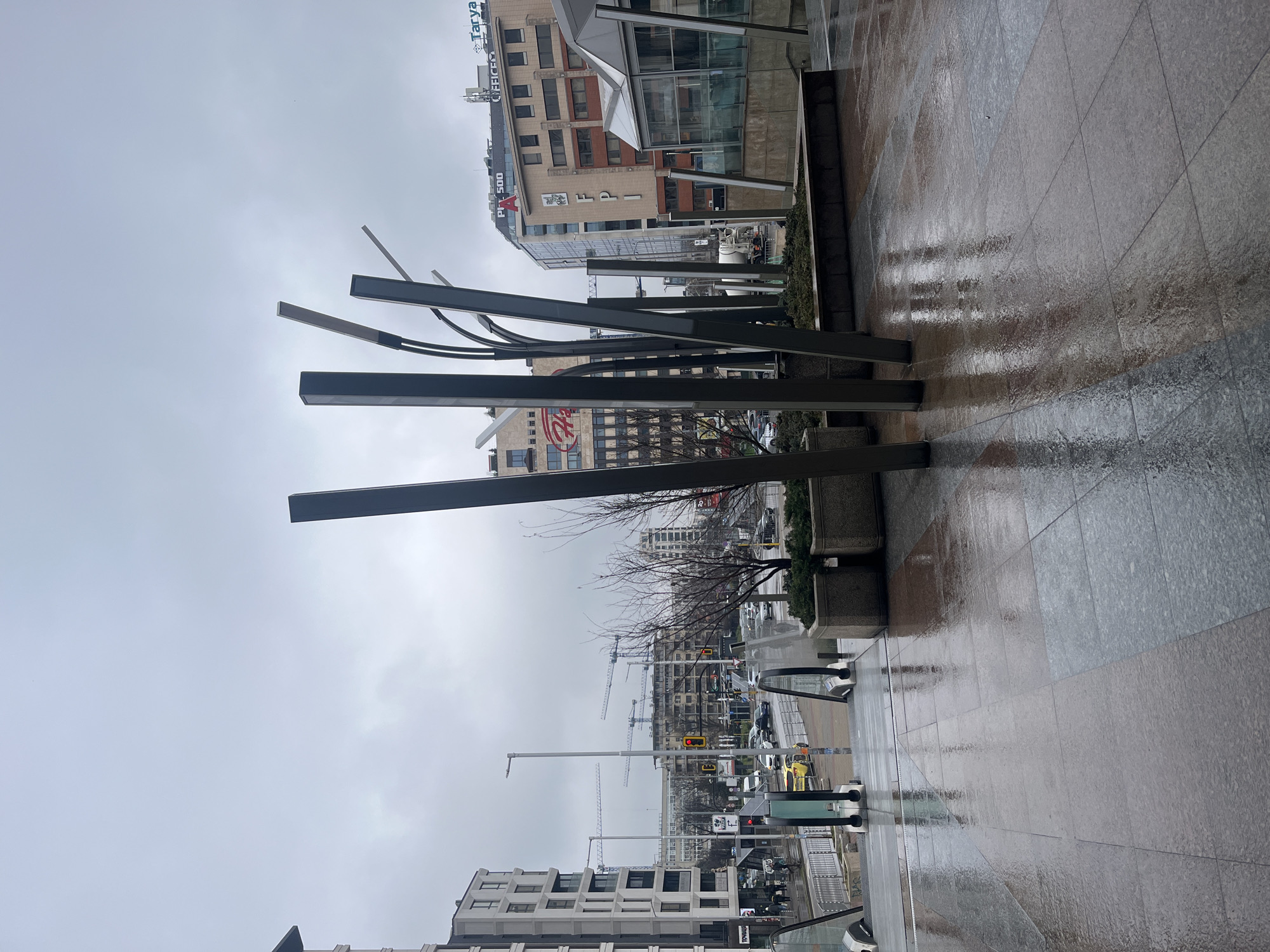 The image depicts an overcast, damp urban environment in Sofia, Bulgaria, specifically near the Serdika Metro Station plaza, looking towards the intersection of bul. Vitosha and bul. Aleksandar Stamboliyski.
The foreground is a wet, tiled pedestrian plaza reflecting the grey sky and surrounding structures. Dominating the immediate foreground is a contemporary public art installation consisting of several dark, slender, angled vertical pillars, some featuring linear light fixtures, emerging from a raised planter box containing low green shrubbery. To the right, glass and metal balustrades delineate a pair of escalators descending into a metro station entrance. Another concrete planter box with greenery is visible on the right.
The midground reveals a multi-lane city street with vehicular traffic, including several passenger cars and a yellow taxi, a white concrete mixer truck, and a red traffic signal. Pedestrians are sparsely visible on the sidewalks. Streetlights line the thoroughfare. A modern, multi-story building with a grid-patterned facade and ground-level commercial establishments is prominent on the left.
The background features various multi-story commercial and residential buildings under a uniformly grey, overcast sky. Prominent building signs include "FPI," "PI 500," "Tarya," and "OFFICE C." Multiple tower cranes are visible in the distance, indicating ongoing construction activity within the urban landscape. The overall scene suggests a cool, wet day with subdued natural light.