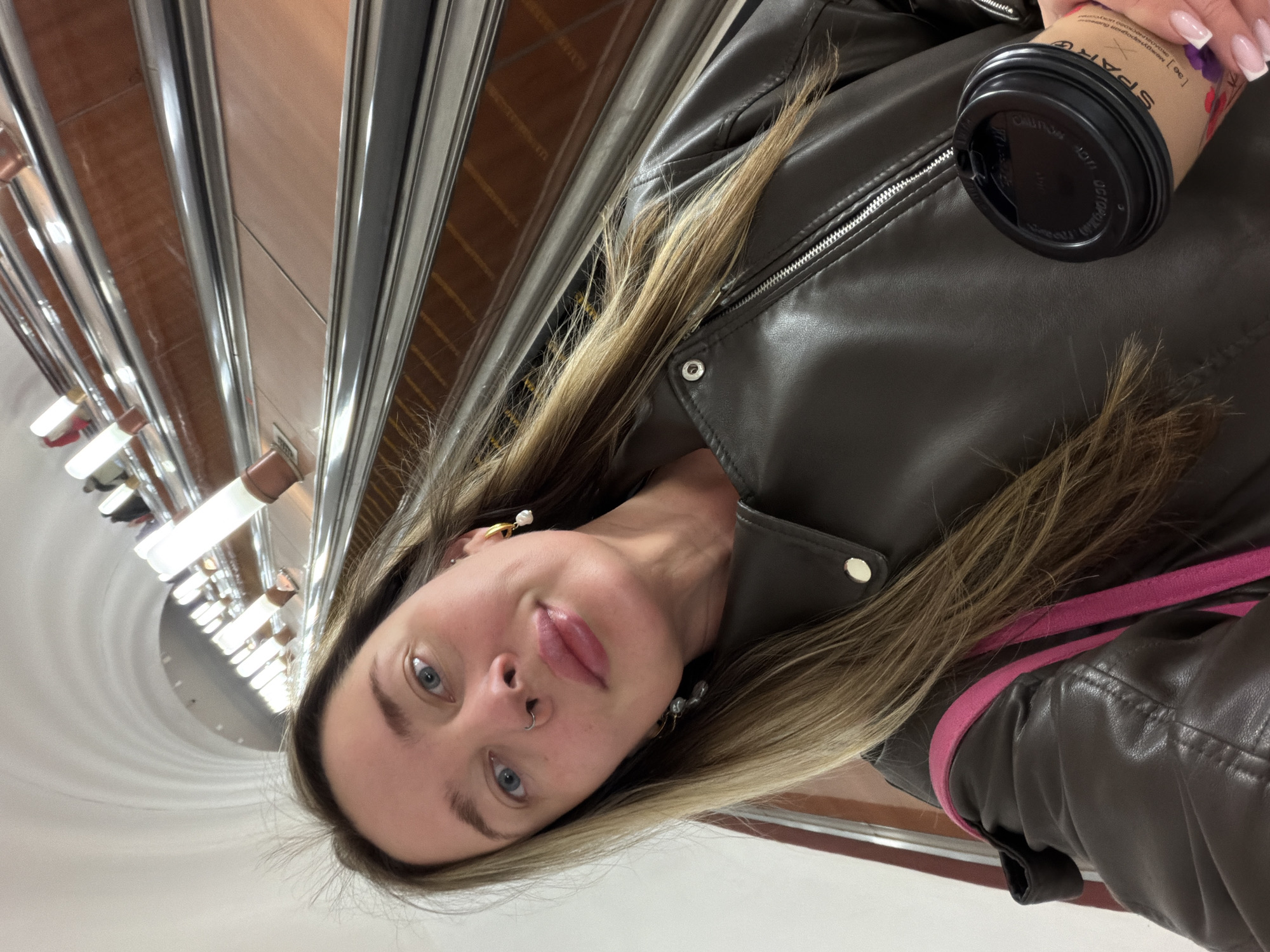A woman with light blonde hair and blue eyes is in the Moscow Metro. She wears a brown leather jacket and gold hoop earrings. She holds a brown paper coffee cup with a black lid. The background shows the metro station's architectural details, including the ceiling lights and tiled walls. The escalator handrails are visible.