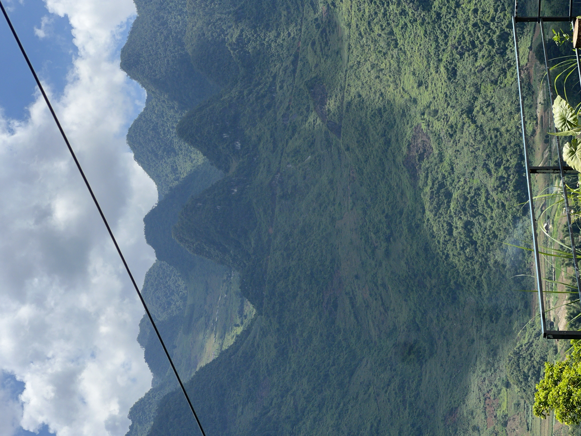 This is a vertical, eye-level, full shot of the mountainous landscape of Lùng Tám Commune, Vietnam. The mountains are covered in dense green foliage, with a cable running diagonally across the sky, from the upper left to the lower right corner of the image. The sky above is partly cloudy, with patches of blue sky visible. In the foreground, a metal railing can be seen, with some green plants growing around it. Below the railing, a valley with terraced fields is visible, with a few small buildings nestled among the greenery. The overall impression is one of a lush, verdant, and remote mountain region.