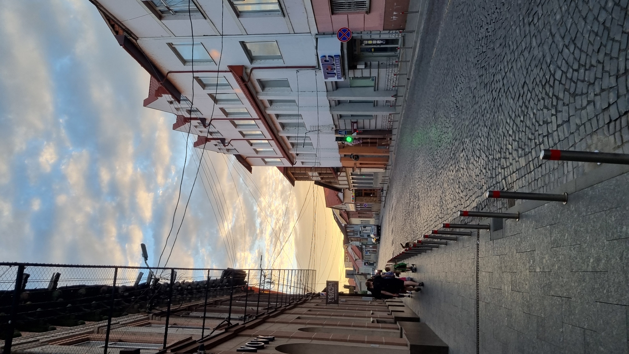 The image depicts a street scene in Mukachevo, Ukraine, at dusk. The street is paved with cobblestones and lined with buildings on both sides. The sky is filled with dramatic clouds illuminated by the setting sun.
On the left side of the street, a building with a light-colored facade and arched windows is visible. A sign with Cyrillic lettering hangs from this building. Further down the sidewalk, several people are walking, including adults and a child pulling a small cart. Metal bollards with red tops line the edge of the sidewalk.
On the right side of the street, buildings with multiple floors and red-tiled roofs are present. One building features a sign with Cyrillic text, and a traffic light shows a green signal. Further down, another building has pink-colored walls and barred windows. The street is relatively empty of vehicles, with one car visible in the distance. The overall atmosphere is serene and captures the characteristic architecture of a European city at the end of the day.