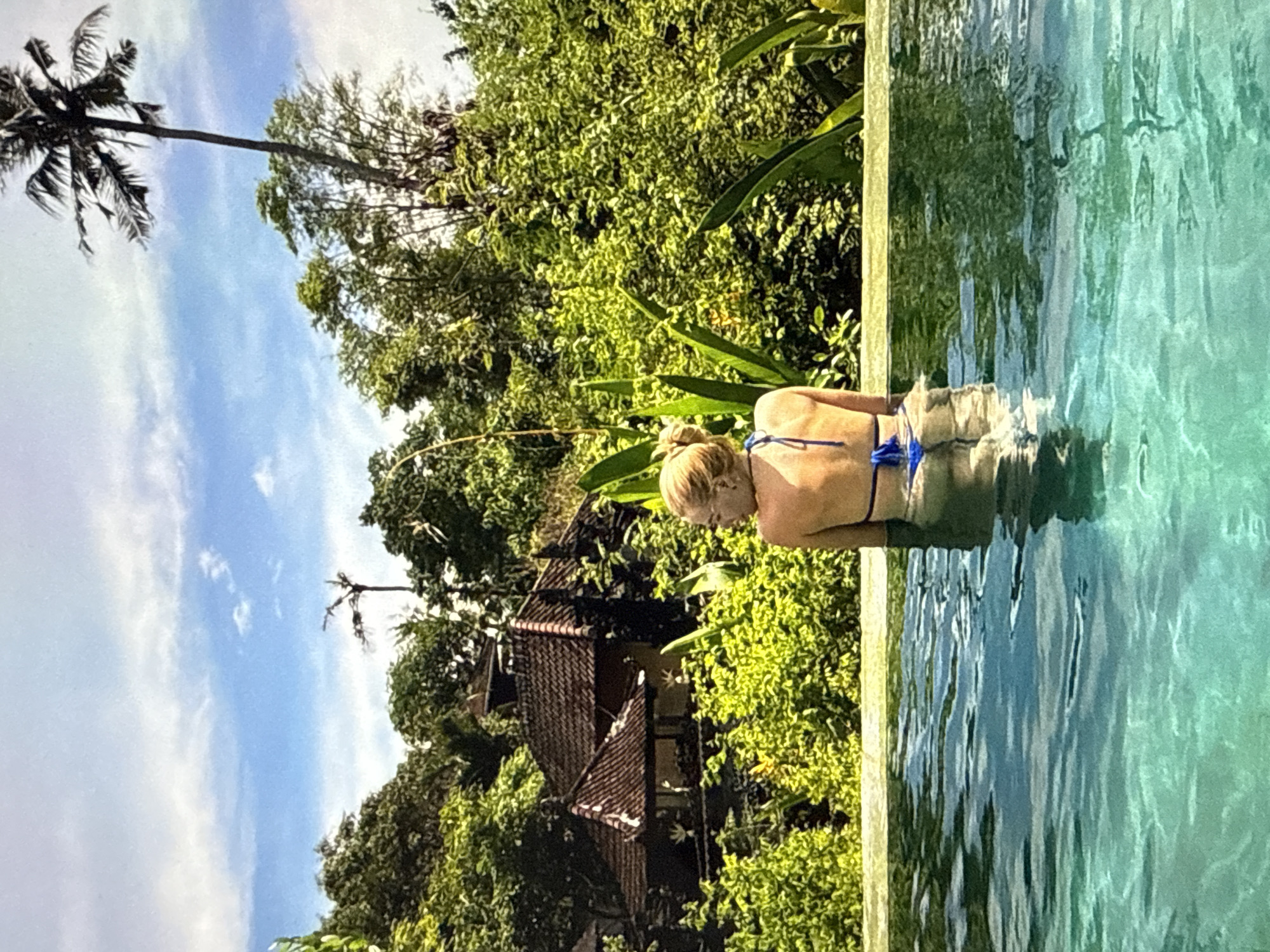 A woman with blonde hair pulled into a bun is seen from behind, seated in an infinity pool overlooking a lush, tropical landscape. She is wearing a blue bikini. The pool water is a clear turquoise with light reflecting off its surface. Behind her, a dense jungle canopy is visible, with a traditional-looking building with a tiled roof nestled amongst the trees. A tall palm tree stands out against a bright blue sky with scattered white clouds. The overall scene suggests a serene and exotic vacation setting.