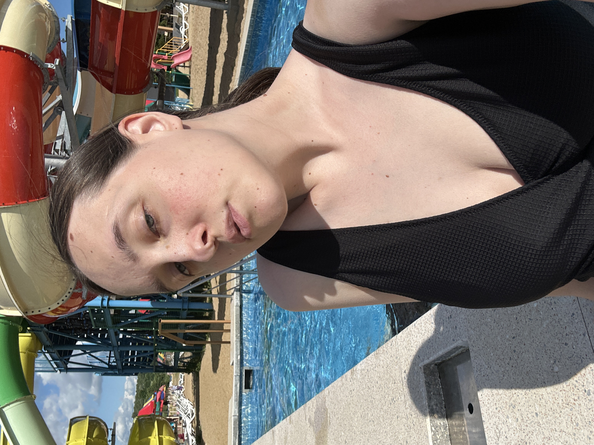 A young woman is posing for a selfie at a water park in Yubileyny, Russia. She is wearing a black swimsuit and is standing next to a pool. In the background, there are several water slides and lounge chairs. The sky is blue and there are some clouds. The woman has light skin and dark hair. She is looking at the camera with a neutral expression.