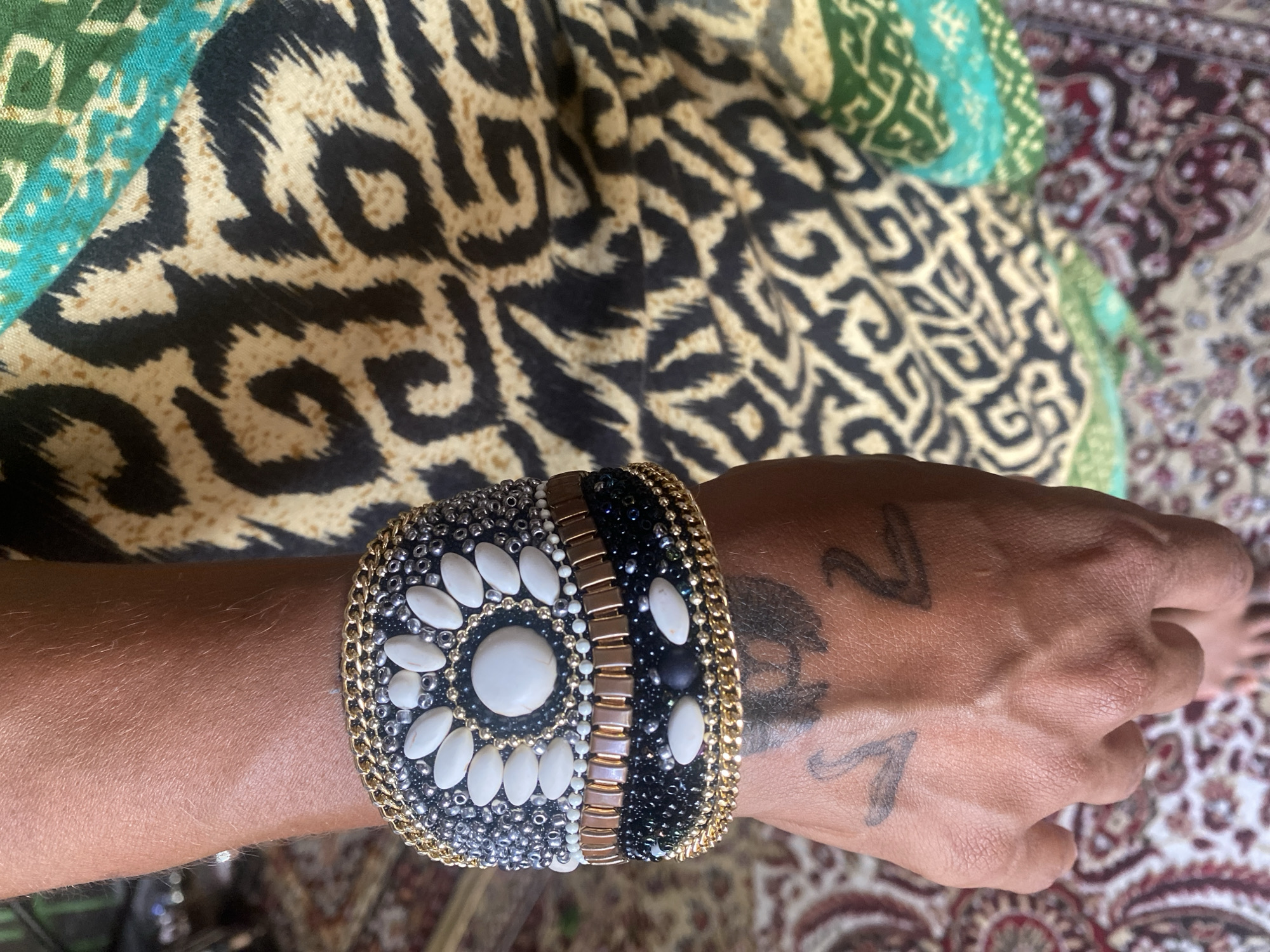 A high-angle close-up depicts a person's left forearm and hand, adorned with a decorative, wide bracelet, set against backgrounds of patterned fabric and carpeting. The bracelet features a black beaded base, white cabochon stones arranged in a circular pattern around a central larger stone, and gold chain edging. The person's skin exhibits visible veins and two small, stylized tattoos. The background includes a swatch of fabric with a black and beige animal print and another section of patterned fabric with green and blue tones. A portion of a multi-colored oriental rug is visible in the lower right corner of the frame. No specific information indicates this is Baan Nai Suan, Thailand.