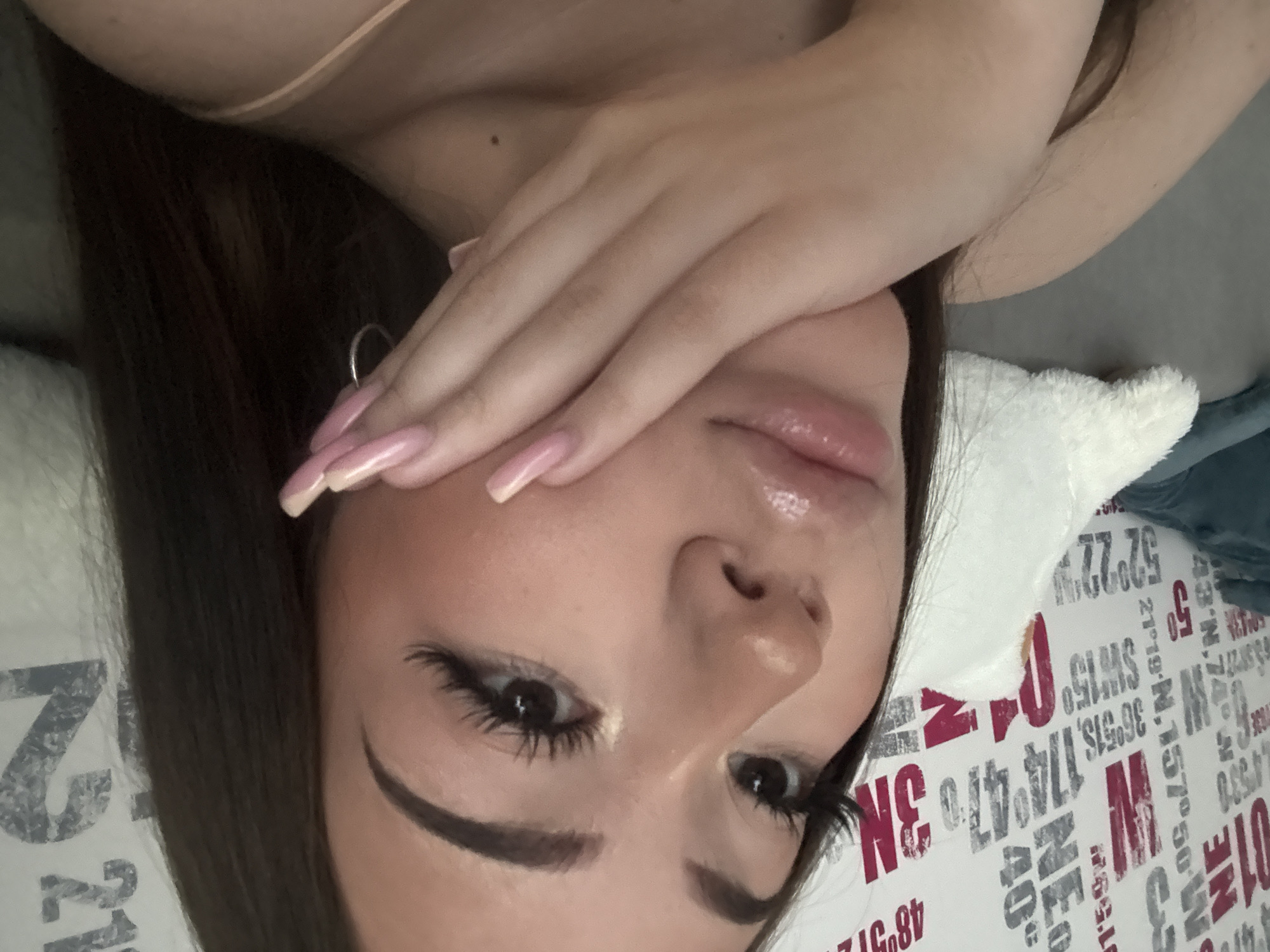 A close-up, angled selfie depicts a woman with dark hair and fair skin lying on a pillow. Her left hand partially covers her face, showcasing long, pink-painted nails. She has prominent eyebrows, long eyelashes enhanced with mascara, and full lips. The pillow beneath her head is printed with coordinates and lettering in red and grey. The limited visible background suggests an indoor setting. The image does not provide enough information to locate it in Naberezhnye Chelny, Russia.