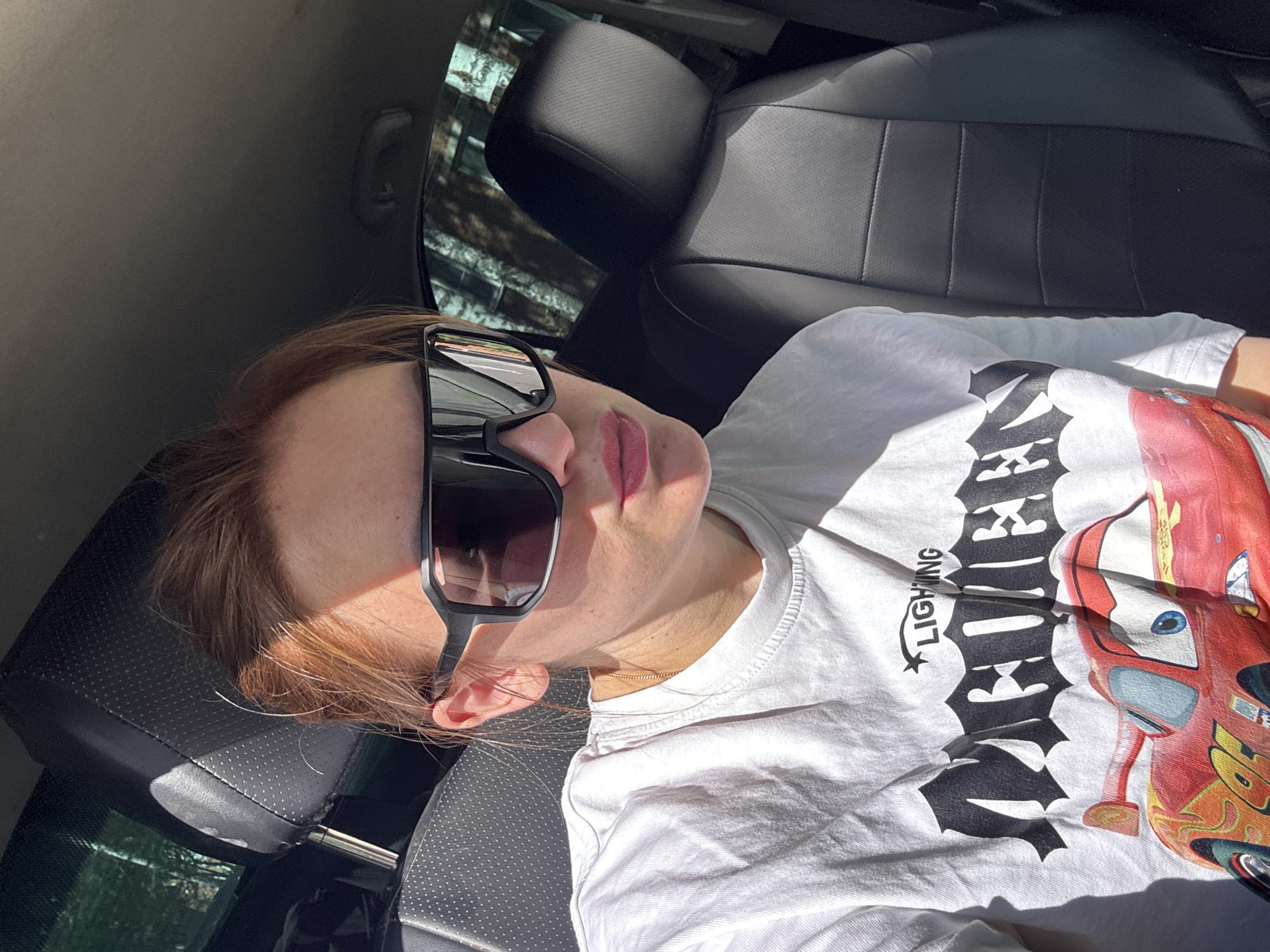 A single individual, appearing female, is seated in the driver's seat of a vehicle in the city of Noviy, Russia. She has light reddish-brown hair pulled back, and wears large, black-framed, reflective sunglasses that obscure her eyes. Her lips are tinted with pink lipstick. A thin gold chain is visible around her neck. She is wearing a white short-sleeved t-shirt featuring a black "LIGHTNING MCQUEEN" graphic in a gothic-style font, with a cartoon depiction of the red and yellow character Lightning McQueen below it. Strong, direct sunlight illuminates the left side of her face and shirt, casting distinct shadows across her right cheek and chin. The car's interior consists of black leather seats and headrests; the driver's headrest, with perforated detailing and chrome supports, is visible behind her. The empty passenger seat, also black leather, is visible to her right. The car's ceiling is dark gray, with a grab handle visible on the passenger side. Through the tinted windows, blurred green foliage and trees are discernible, indicating an outdoor, daytime setting. No specific action is depicted beyond sitting and facing forward.