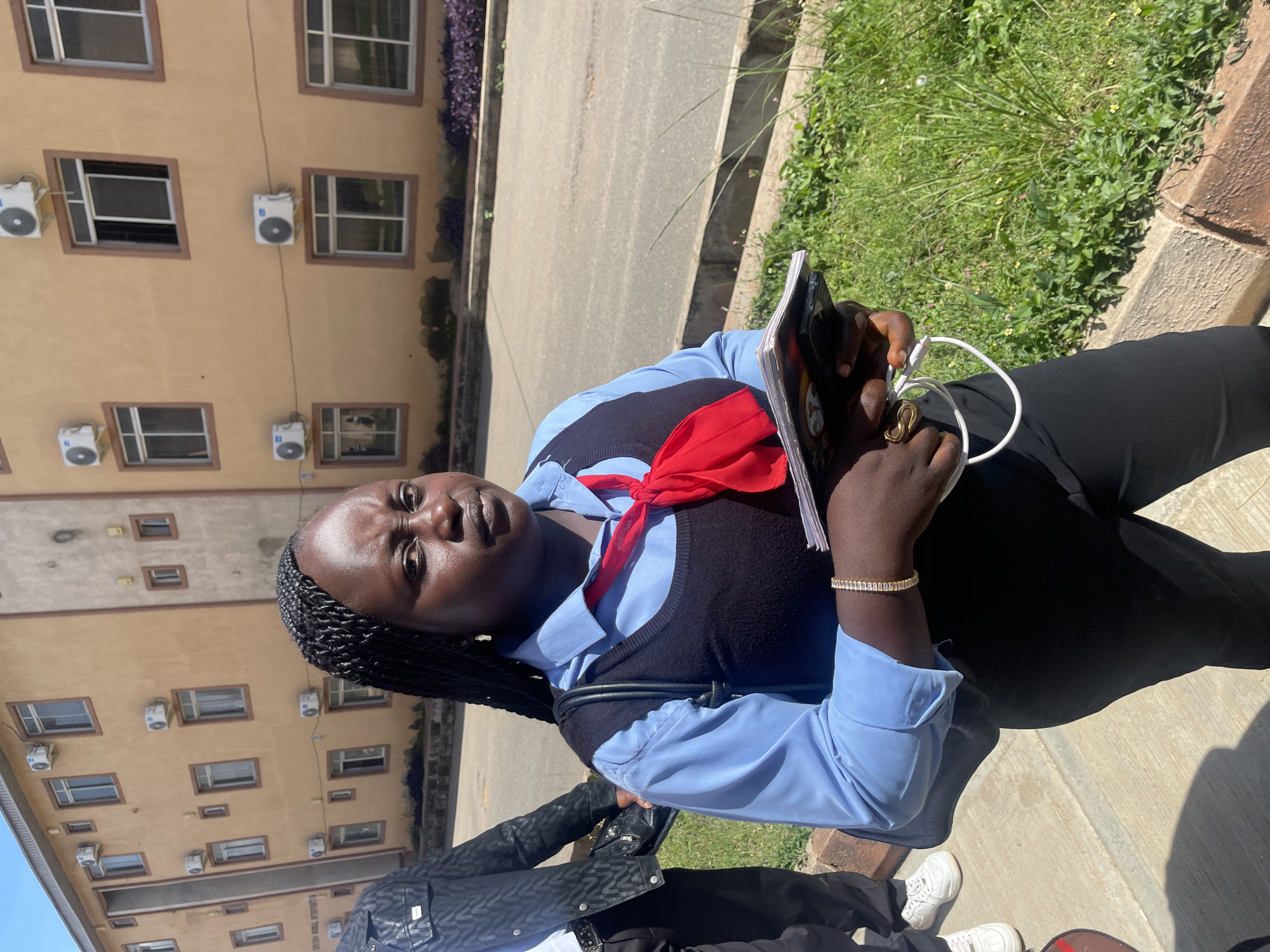 A dark-skinned woman with braided hair and a red scarf is standing in front of a tan building. She is wearing a light blue shirt, a black vest, and black pants. She is holding a notebook and a white cord. The building has several windows and air conditioning units. The woman is looking at the camera with a serious expression. The ground is paved, and there is grass and a curb on the right side of the image. The image appears to be taken outdoors in Rafin Par, Nigeria.