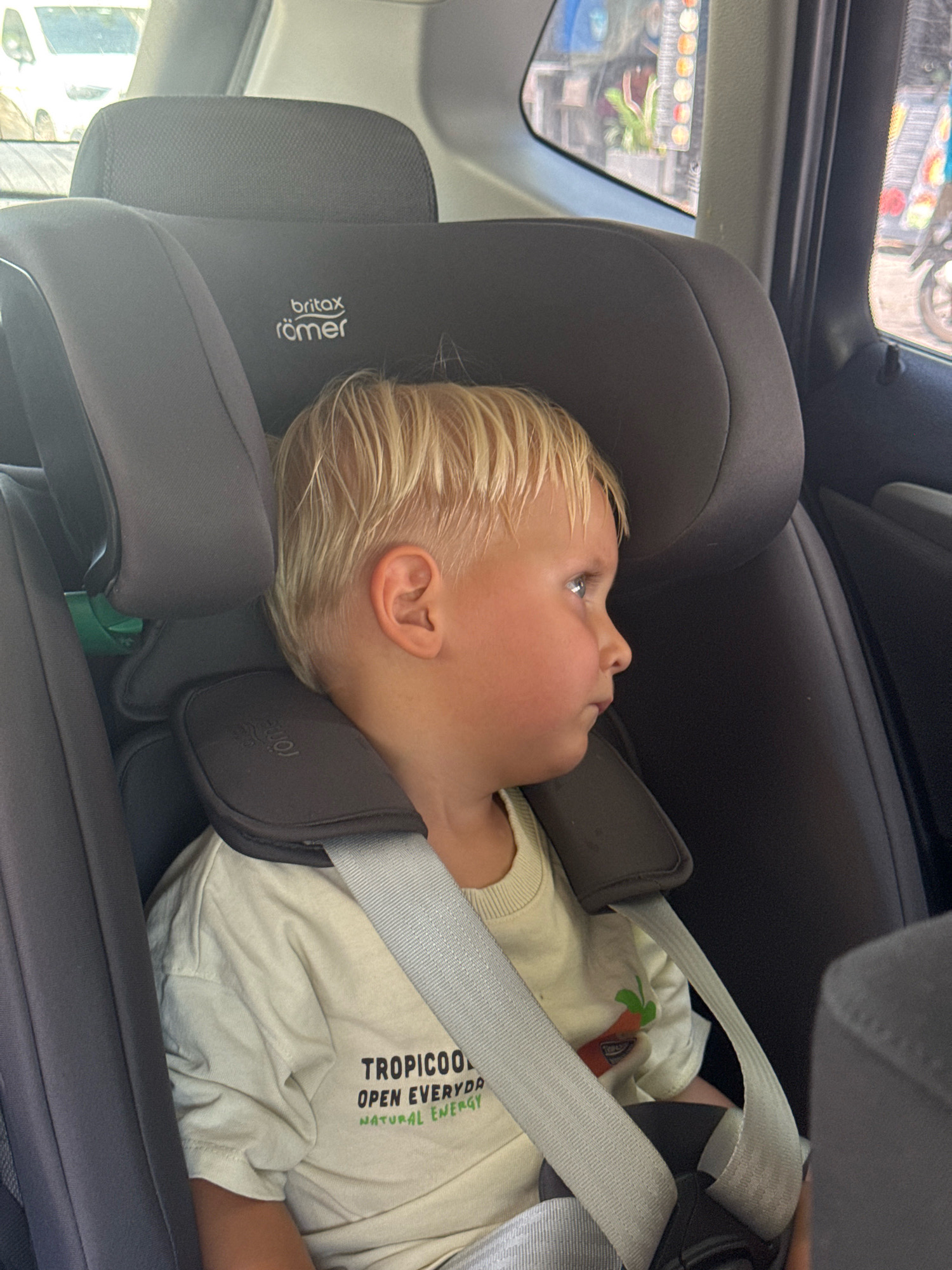 A young blonde-haired boy is seated in a gray car seat, looking out the window to his right. The boy wears a light-colored t-shirt with black lettering that reads "TROPICOOL OPEN EVERYDA NATURAL ENERGY" and a small graphic of a carrot. A gray seatbelt is fastened across his chest. The car appears to be in motion, as suggested by the blurred view of the outside environment through the car window. The context of Germasogeia Municipality, Cyprus, places this scene within a Mediterranean urban or suburban setting.