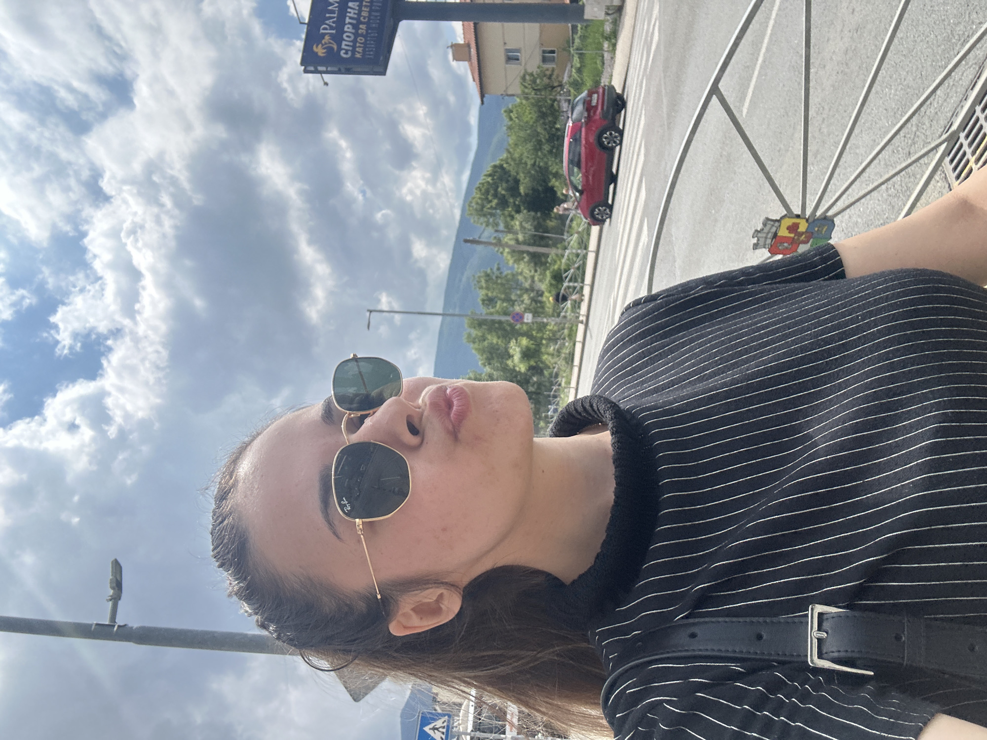 This image is a vertical selfie, rotated 90 degrees clockwise, taken outdoors in Sofia, Bulgaria. The main subject is a young woman, positioned in the foreground. She is looking upwards and to the right, with her lips in a pouting expression. She wears dark, hexagonal Ray-Ban sunglasses and a black top with fine white horizontal stripes, along with a black belt featuring a silver buckle.
The background reveals a street scene under a bright, partly cloudy sky. A red car is visible on a pedestrian crossing (zebra lines). Further in the distance, green trees, buildings, and large mountains are discernible, indicating an urban setting nestled against natural scenery. A blue billboard with Cyrillic text is prominent in the upper left. The visible text reads "PALMA СПОРТНА" (Palma Sportna) and partially "КАТО ЗА СВЕТО..." (Kato za Sveto...). A decorative, colorful, shield-like object is attached to a metal fence or railing in the mid-ground.
The photo captures a casual, playful moment during the daytime. The cloudy sky suggests typical weather for an outing. The Cyrillic text and the mountainous landscape are key location cues, consistent with Sofia, Bulgaria.