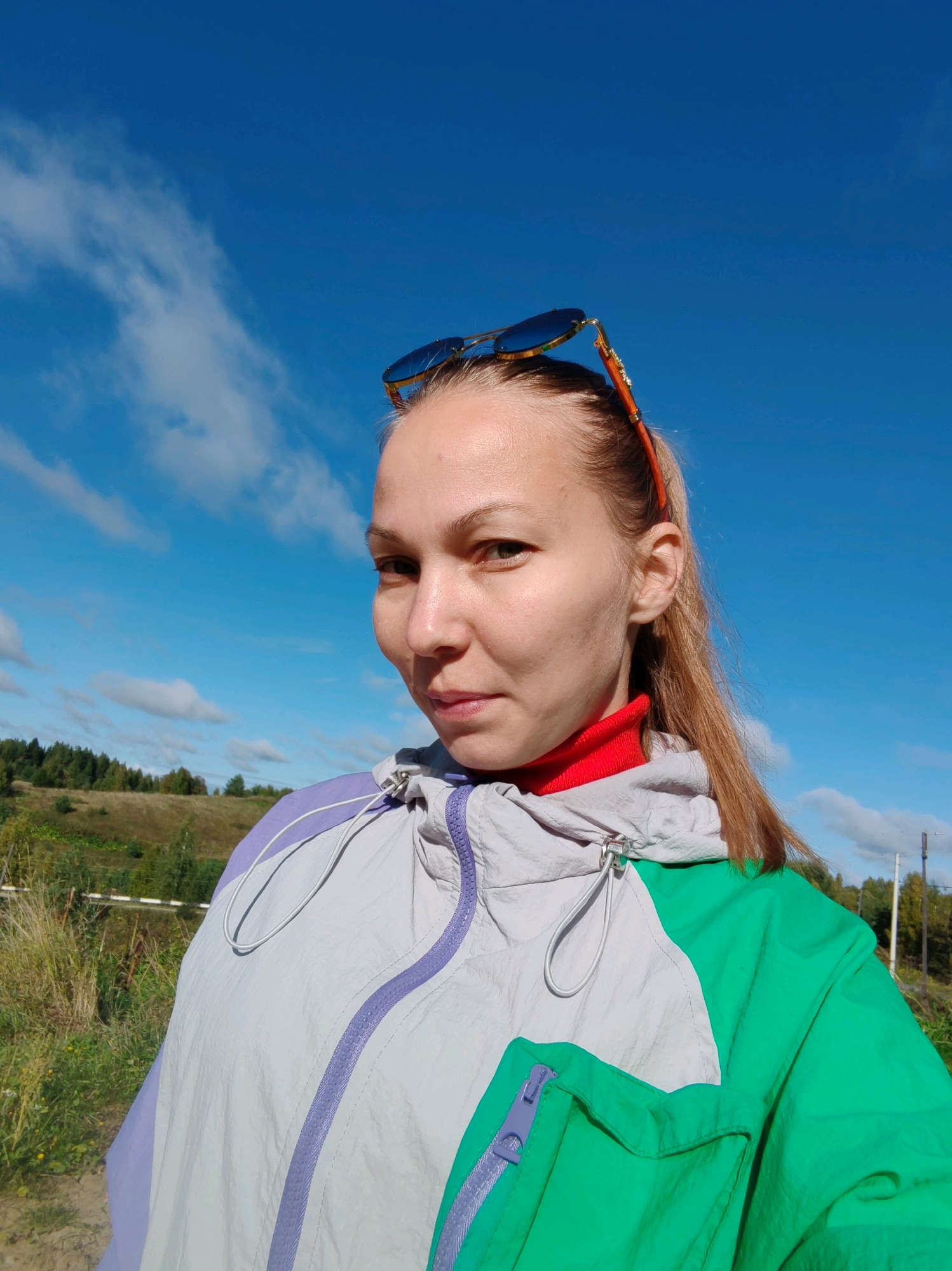 A close-up, waist-up, selfie-style, outdoor portrait of a Caucasian woman with blonde hair pulled back and sunglasses perched on her head. She is wearing a green and grey hooded jacket with purple accents and a red turtleneck shirt. The woman is looking directly at the camera. The background shows a blue sky with scattered clouds and a grassy, wooded landscape. This scene is located in Chukhlem, Russia.