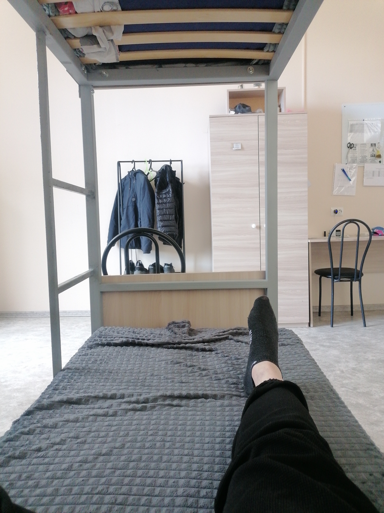 The image depicts a room that appears to be a dormitory or a small apartment. In the foreground, a person's legs, clad in black pants and socks, are resting on a grey, textured blanket spread across a bed. The bed is part of a lofted bed frame, with the lower bunk visible, also covered in a similar grey blanket. Above the bed, laundry is stored on wooden slats.
The background reveals a functional living space. To the left, a black metal clothing rack holds two jackets, a dark blue one and a black puffer jacket, along with what appear to be shoes placed below. Further to the right, a light wood wardrobe stands against the wall. Adjacent to the wardrobe is a white desk with a black chair, and on the wall above the desk are several papers and notices pinned to a noticeboard, along with an electrical outlet and a pen. The walls are a neutral cream color, and the floor is a light grey carpet.
The overall impression is of a tidy, yet lived-in personal space. The specific location of Комсомольский, Russia, is mentioned in the prompt but cannot be visually verified from the image itself.