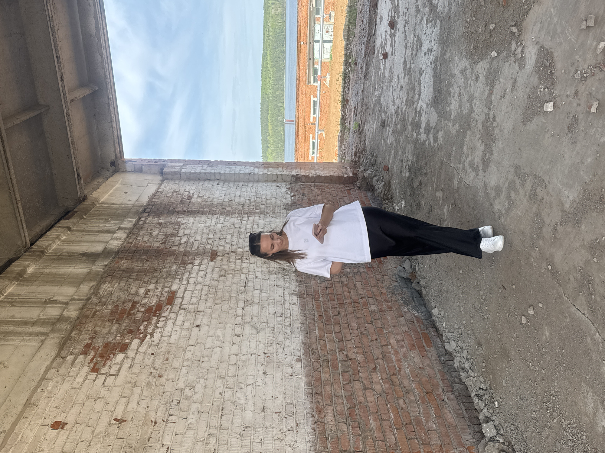 The image depicts a person standing within the ruined structure of a building. The person is wearing a white t-shirt, black pants, and white shoes. The interior of the building is unfinished with exposed brick walls and a concrete floor. Through an opening in the structure, a lake and buildings in the distance are visible. Based on visual clues, the location is potentially Chaikovsky, Russia.