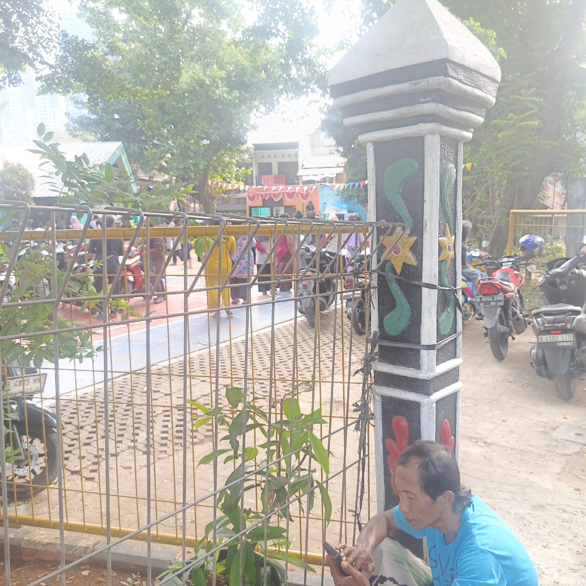 The image depicts an outdoor scene with a focus on a decorative pillar in the foreground, adorned with painted green floral motifs and yellow stars. Behind the pillar, a metal fence separates the viewer from a paved area where several motorbikes are parked. In the background, a group of people, mostly women wearing headscarves and traditional attire, are visible, some walking and some gathered around the motorbikes. A building with a colorful banner is also discernible. To the far left, a tall building, possibly a skyscraper, is partially visible against a bright sky. A man in a blue t-shirt is seated in the lower right corner of the frame, looking at his mobile phone. The ground appears to be a mix of sand and paving stones. The overall impression is of a public outdoor space, possibly a schoolyard or community area, with parked motorbikes suggesting a mode of transportation in use. The presence of a skyscraper in the background hints at an urban setting within South Jakarta, Indonesia.