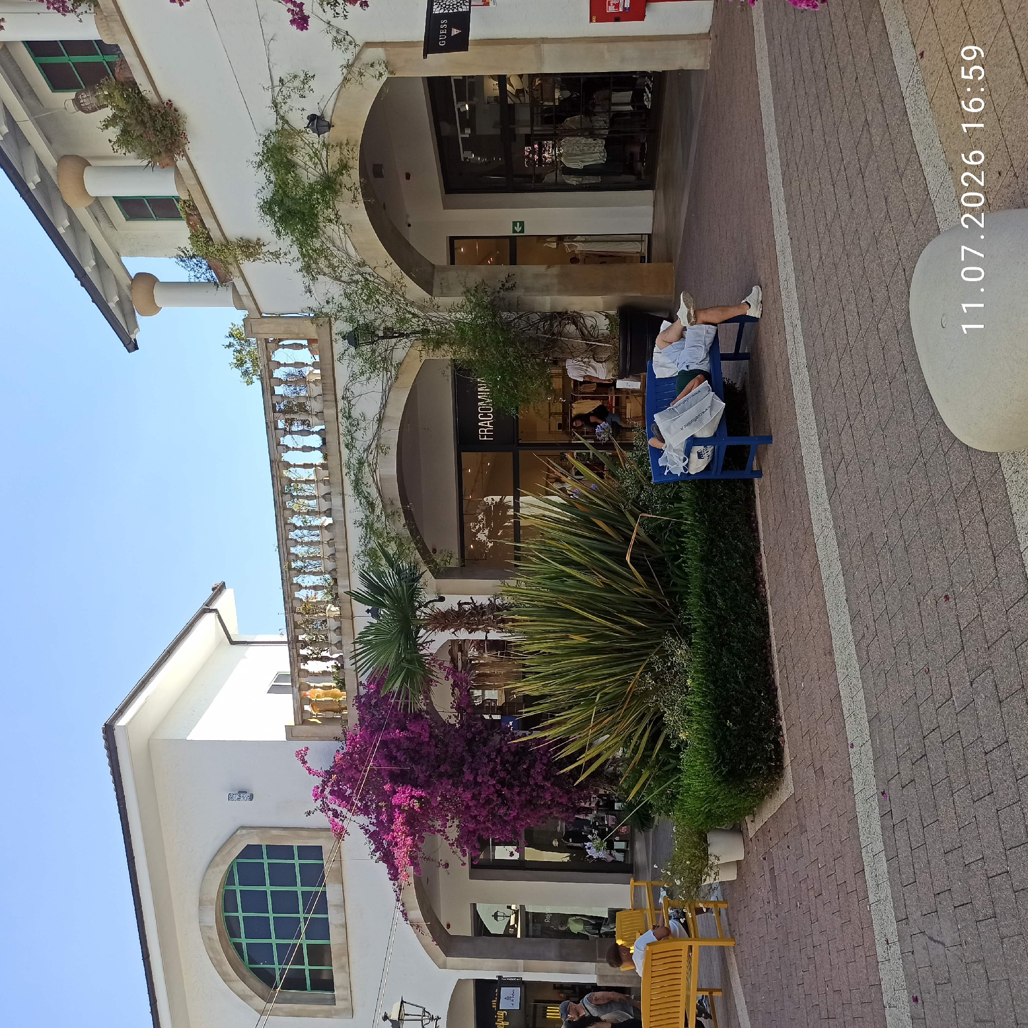 In Molfetta, Italy, at an outdoor shopping area, a person is resting on a blue bench, covered by a white shopping bag. The bench is situated on a paved walkway with brick patterns and a white stripe. To the left, a yellow bench is occupied by two individuals, one of whom is looking at their phone. In the background, there are white buildings with arched doorways, storefronts, and lush green foliage, including a large bougainvillea plant with vibrant pink flowers. The sky above is clear and blue. The timestamp "11.07.2026 16:59" is visible in the lower right corner of the image.