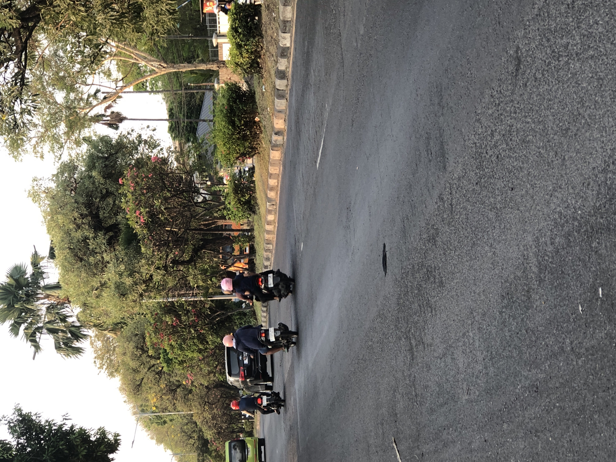 The image shows a street scene in the city of Ngaban, Indonesia. Three individuals are riding motorcycles on a paved road. The person on the left is wearing a blue shirt and a pink helmet, riding a black motorcycle. In the middle, another person in a dark jacket and pink helmet is also on a black motorcycle. To the right, a third individual, wearing a dark shirt and pink helmet, is riding a black motorcycle. A black SUV is visible behind the middle and left motorcyclists. A green bus is parked on the left side of the road further down. The environment includes lush green trees and bushes lining the street, with a clear sky above. The road has a white line marking a lane. The sun appears to be low in the sky, casting long shadows.