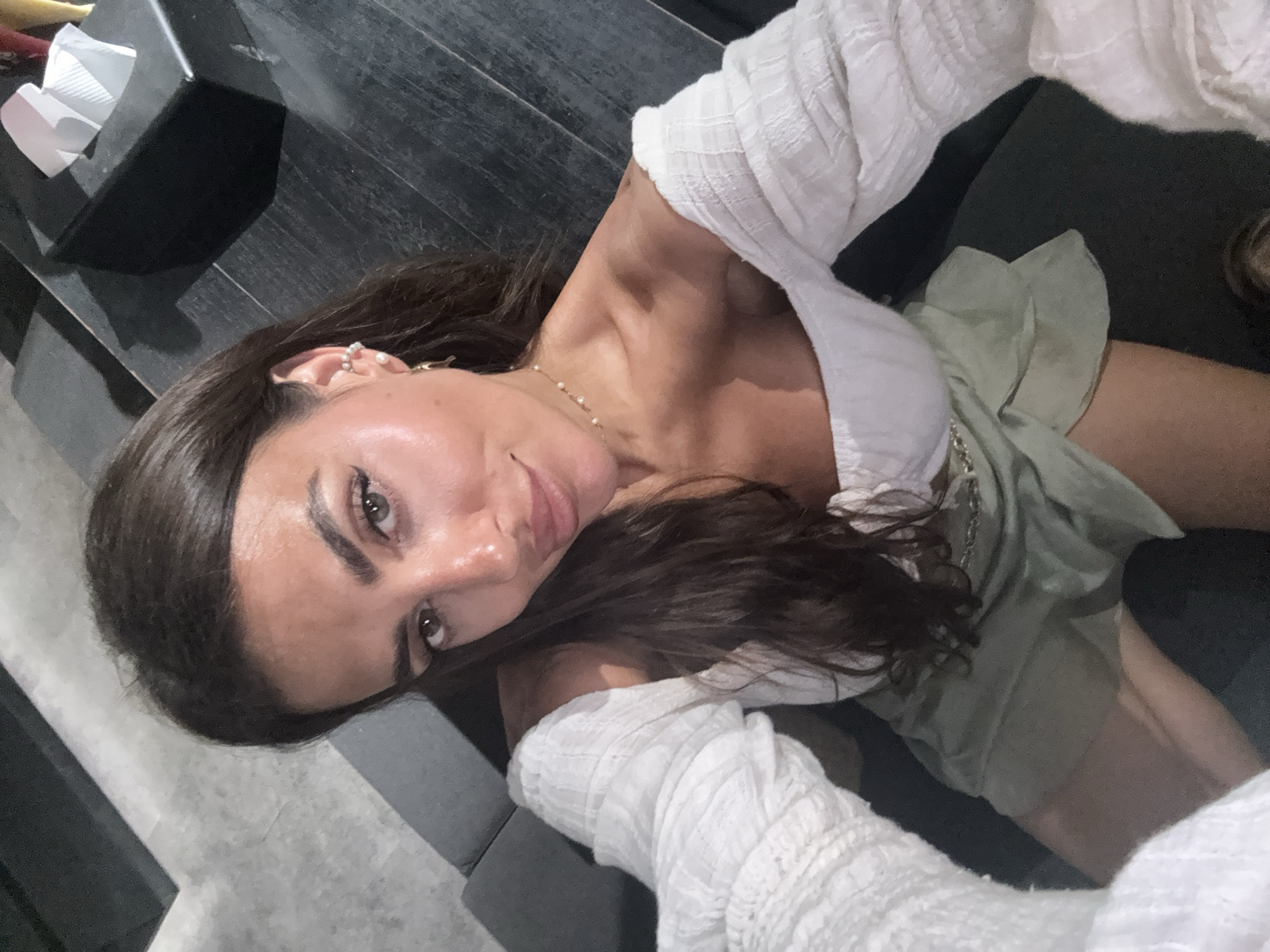 A woman with dark brown hair and olive skin is taking a selfie. She is wearing a white, off-the-shoulder top with textured sleeves and a light green skirt or shorts. She is wearing a pearl necklace and multiple earrings on her left ear. To the upper left of the frame is a black, rectangular box of tissues. The woman appears to be sitting on dark furniture, possibly a couch. The background includes a light gray floor. The location is reported as North Kuta, Indonesia.