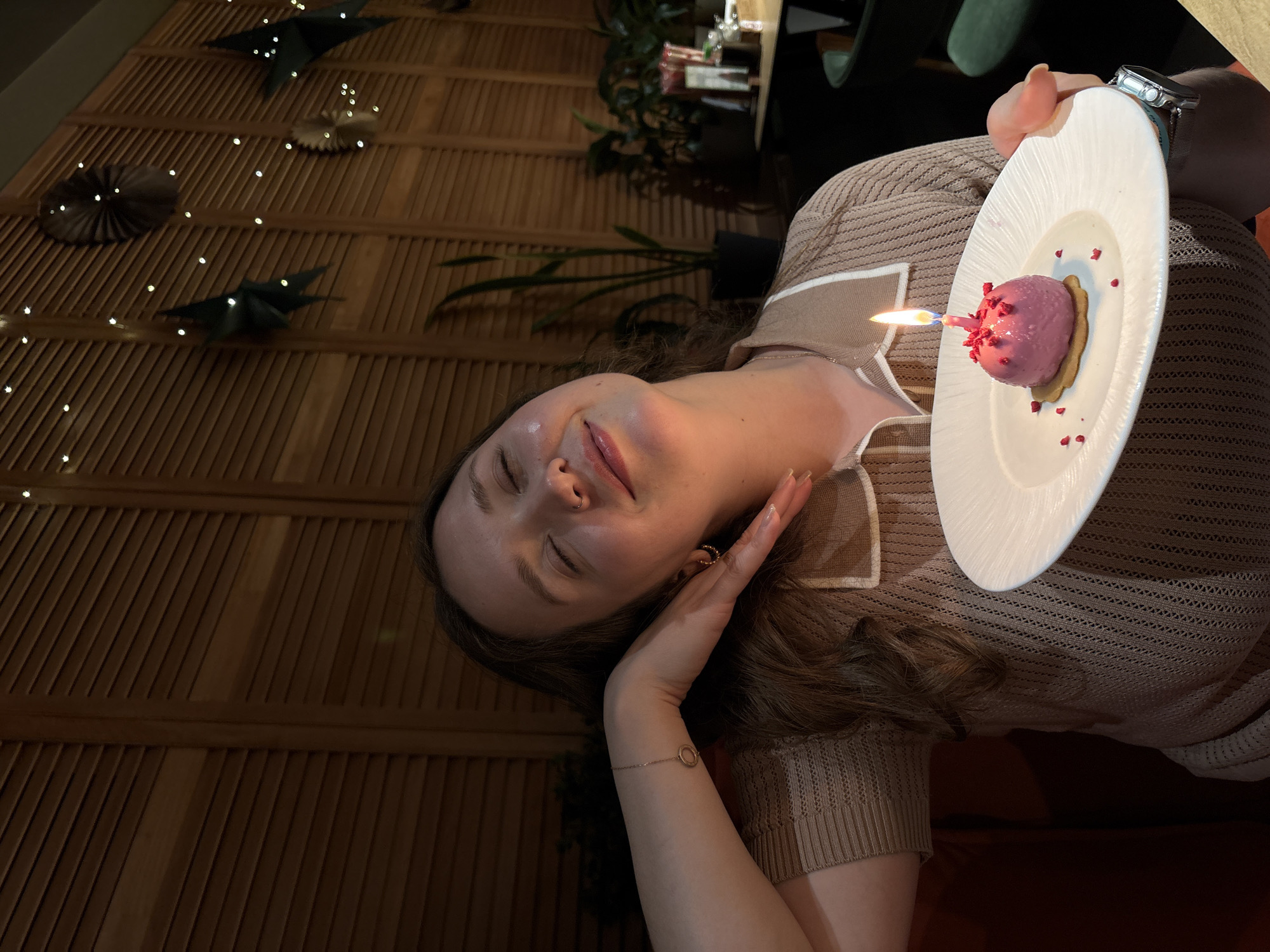 The image captures a young woman celebrating in what appears to be a dimly lit restaurant or cafe in Izhevsk, Russia. She is positioned vertically, her face turned slightly to the right with her eyes gently closed and a soft, happy smile, suggesting a moment of contentment or making a wish. She has light skin, dark wavy hair, a nose ring, and small hoop earrings.
She is dressed in a light brown or beige knitted top with distinct white trim. Her left hand rests gracefully near her face, revealing a delicate bracelet with a circular charm on her wrist. In her right hand, which also wears a silver watch with a dark strap, she holds a white plate. On the plate rests a small, round, pink dessert, likely a mousse or ice cream bomb, topped with red sprinkles or crumbs and sitting on a cookie or biscuit base. A single, lit birthday candle with a bright flame is stuck into the dessert, clearly indicating a birthday celebration.
The background features a textured slatted wooden wall panel adorned with numerous small, twinkling fairy lights and several dark, folded paper decorations shaped like stars and fans, enhancing the festive and cozy atmosphere. Green plants and blurred dark furniture elements are also visible, further suggesting an indoor dining environment during the evening or nighttime. There is no visible text in the image.