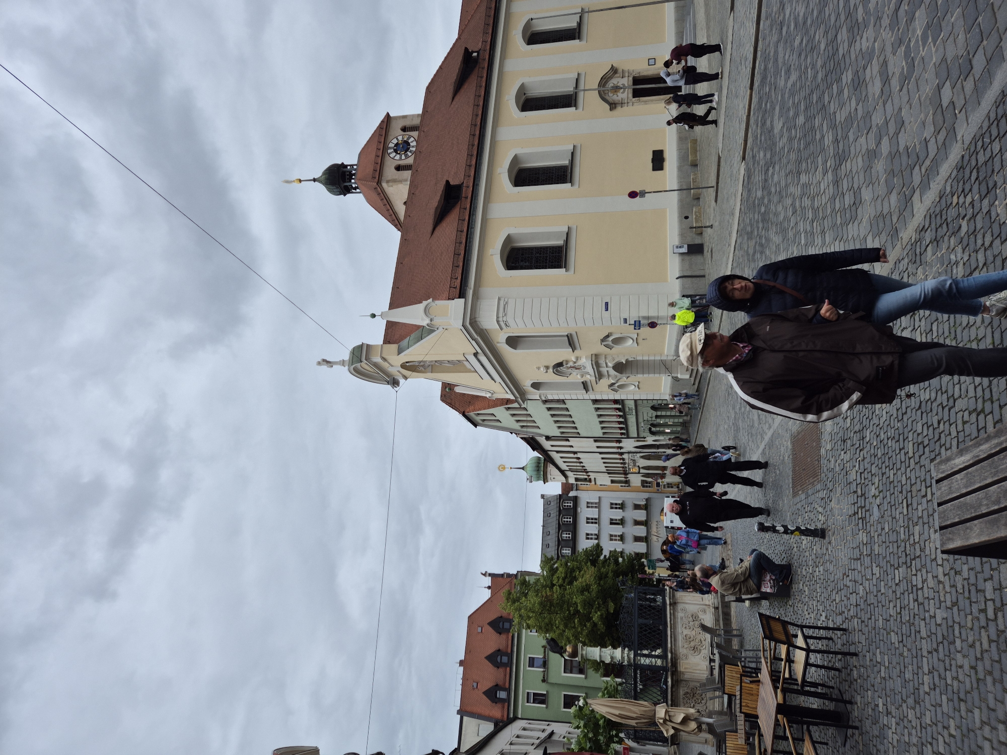 In the city of Regensburg, Germany, a street scene unfolds under a cloudy sky on a cobblestone street. In the foreground, a man wearing a brown jacket and a cap walks with a woman in a dark jacket and jeans. The woman is looking ahead, while the man is looking towards the woman. Behind them, a group of people are walking down the street, some in casual clothing. To the left of the street, outdoor seating for a cafe is visible with tables and chairs. Further back, a fountain stands with a statue on top. Buildings line the street, some with colorful facades and tiled roofs. A church with a prominent clock tower is visible in the background. People are engaged in various activities, such as walking, sitting, and conversing. The overall atmosphere is that of a typical European city street on a slightly overcast day.