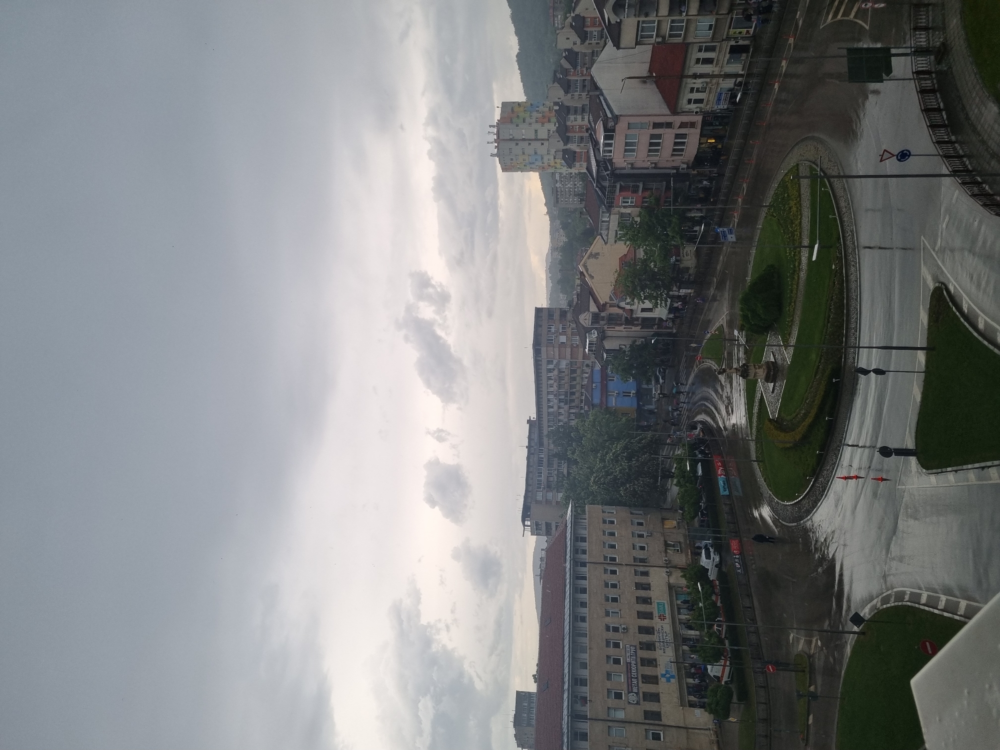 This is an eye-level, outdoor shot of the city of Gabrovo, Bulgaria, during a rainy day. The sky is overcast. A roundabout is visible to the right, with a circular grassy area in the center. The road is wet, reflecting the sky. Buildings of varying heights and architectural styles line the road, including apartment buildings with red roofs. Some buildings have faded paint. Vegetation is visible along the road and in the center of the roundabout. A hill or mountain can be seen in the background behind the buildings. The overall tone is gray and overcast.