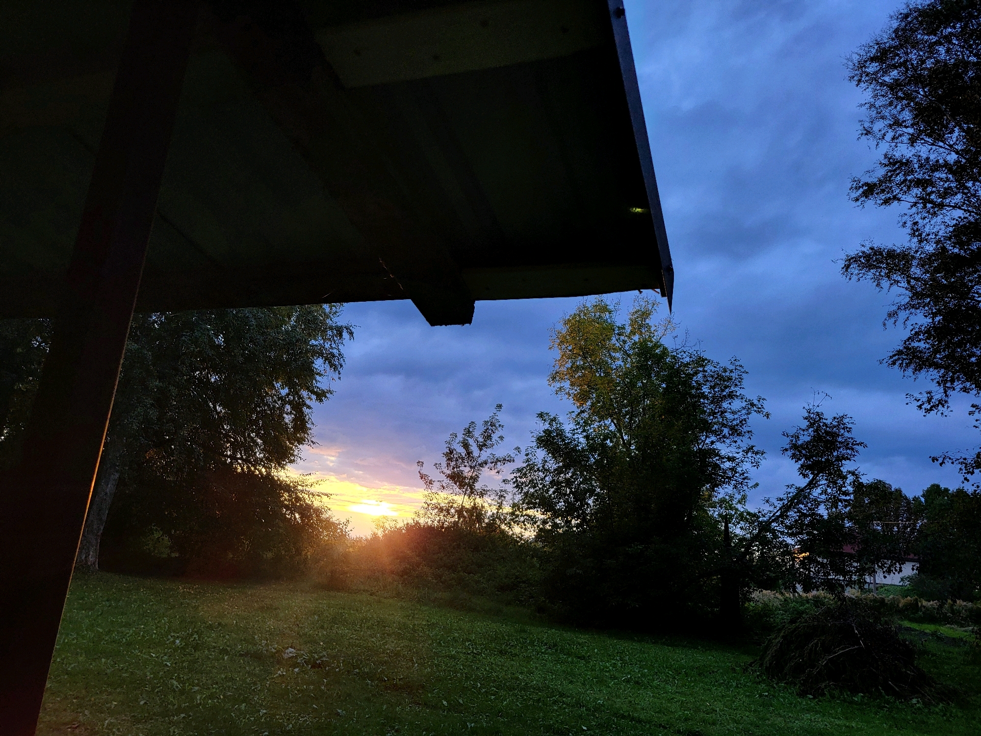 The image depicts a twilight scene in Kalnciems, Latvia. The foreground features a dark, angular structure, possibly a roof or overhang, casting a shadow over a grassy lawn. Beyond the lawn, a line of trees and bushes creates a natural barrier, with the setting sun peeking through the foliage on the left side of the frame, casting a warm glow. The sky is a deep blue, streaked with scattered clouds, indicating the transition from day to night. The overall atmosphere is serene and natural, with no people or immediate signs of human activity beyond the man-made structure.