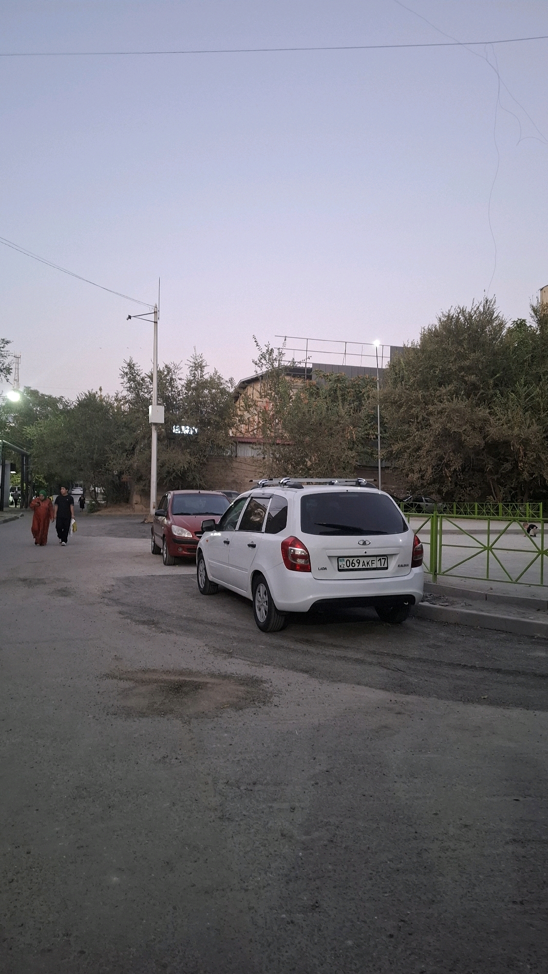 The image shows a street scene in Shymkent, Kazakhstan, during dusk. In the foreground, a white Lada Kalina station wagon is parked facing right. The car's license plate reads "KZ 069 AKF 17". Behind it, a dark red car is partially visible. Two individuals are walking on the left side of the street, one in an orange abaya and the other in dark clothing. A metal fence runs along the right side of the street, separating it from an area with trees and a building in the background. A lamppost with a camera is also visible. The street is paved with asphalt and shows signs of wear and tear, with a visible pothole in the foreground. The sky is a pale gradient of blue and purple, indicating the end of daylight.