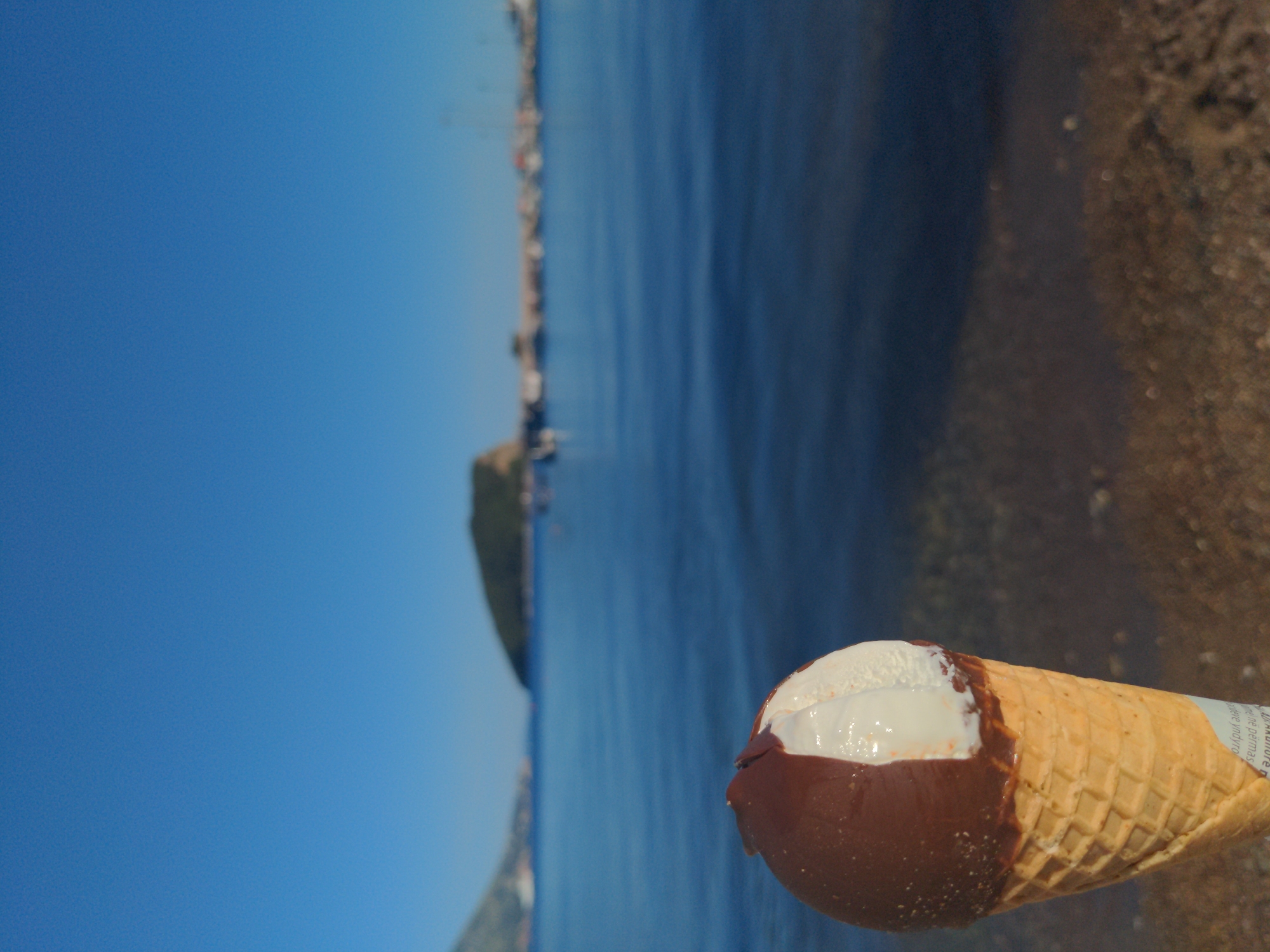 An ice cream cone with chocolate covering the top half of the ice cream is shown, with a waffle cone below. The background appears to be a blue body of water, possibly the ocean. The right of the image shows a sandy beach, and across the water, there is a coastline with buildings and greenery. The location is Unknown, Unknown.
