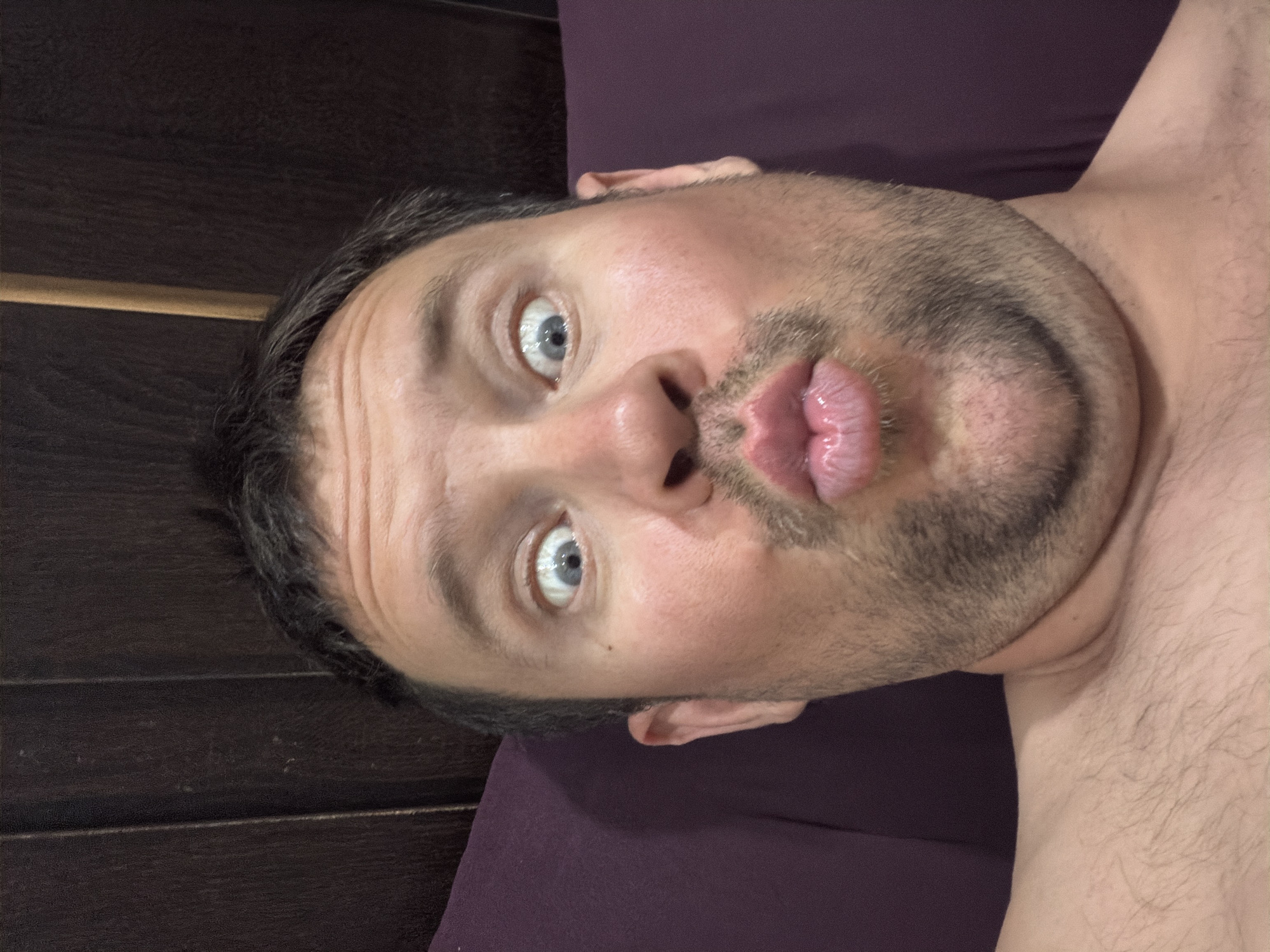 A close-up, selfie-style portrait shows a light-skinned male with short, dark hair and a light beard. He is lying down and looking directly at the camera with his eyes wide open and his lips puckered in a kissing gesture. The man is shirtless, and the background consists of a dark brown wooden headboard and a dark purple upholstered headrest. The image appears to be taken indoors, possibly in a bedroom. There is no indication of the location being Baan Mae Nam, Thailand.