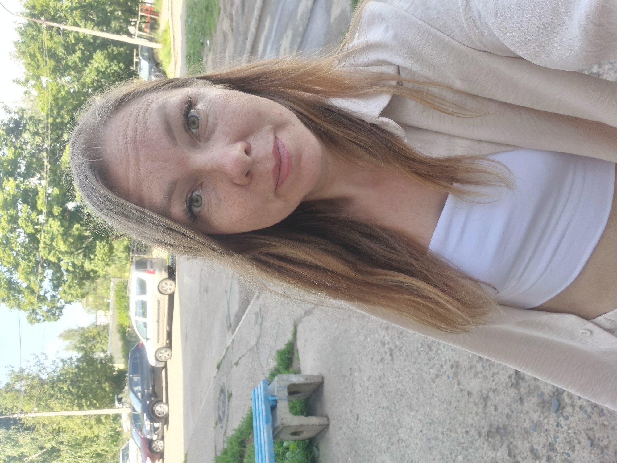 The image is a selfie taken outdoors in Chaikovsky, Russia, on a sunny day. The subject is a woman with shoulder-length blonde hair, green eyes, and freckles. She is wearing a white tube top and an open, cream-colored button-up shirt. In the background, several cars are parked on a paved area, including a black sedan and a light-colored station wagon. To the left, a blue bench with concrete supports is visible on a concrete path, with green grass growing around it. Trees with lush green foliage line the background, and a utility pole is present. The overall environment suggests a residential or public outdoor area within the city.