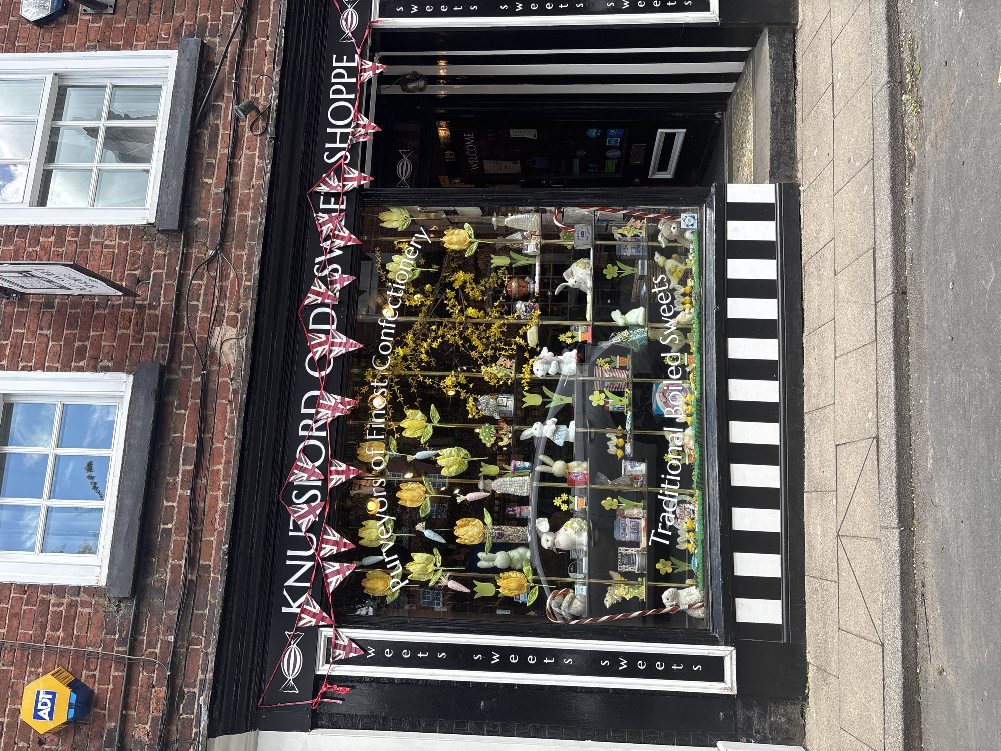 A street-level view presents "Knutsford Olde Sweet Shoppe" in Knutsford, United Kingdom, noted as "Purveyors of Finest Confectionery." The shop facade is black with white lettering. A garland of Union Jack flags stretches across the top of the shopfront. The window display contains an assortment of sweets and floral decorations, including yellow tulips and bunny figurines. Signage within the window reads "Traditional Boiled Sweets." The building's architecture includes brickwork and white-framed windows above the shopfront. An ADT security box is visible on the left side of the building. A portion of the pavement is visible to the right of the shop.