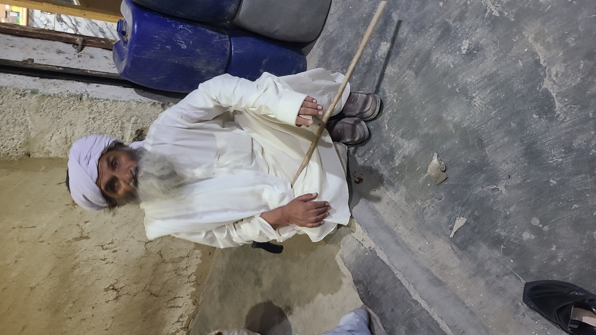 A man with a grey beard and a white turban sits cross-legged on a concrete floor in what appears to be an indoor space. He wears a white outfit and grey shoes. The man holds a wooden stick in his hands, oriented vertically. Behind him are several blue and grey plastic containers. The wall to his left is rough, possibly concrete. The floor also shows signs of wear and discoloration. A black shoe is visible in the lower right corner of the image. The location is Ziarat, Pakistan.