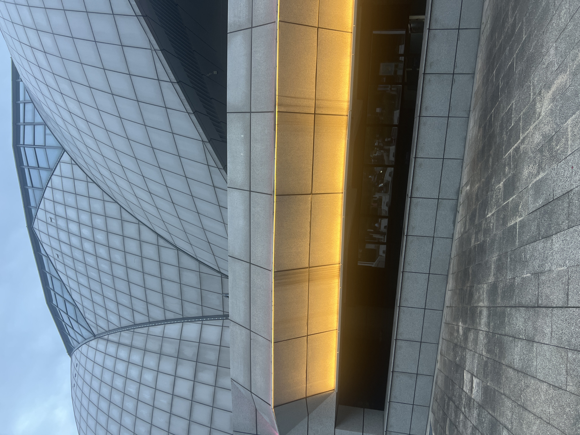 The image shows a modern building facade with a curvilinear roof structure composed of segmented panels, likely glass or a translucent material, under a cloudy sky. Below the roof, a grey, angular section of the building features a horizontal band of warm yellow light emanating from an illuminated recess. This recessed area appears to be a passageway or entrance with dark glass windows reflecting interior lights and figures. The foreground consists of a grey tiled walkway with a visible slope. The location is Liandu District, China. No people are clearly visible or identifiable in the image.