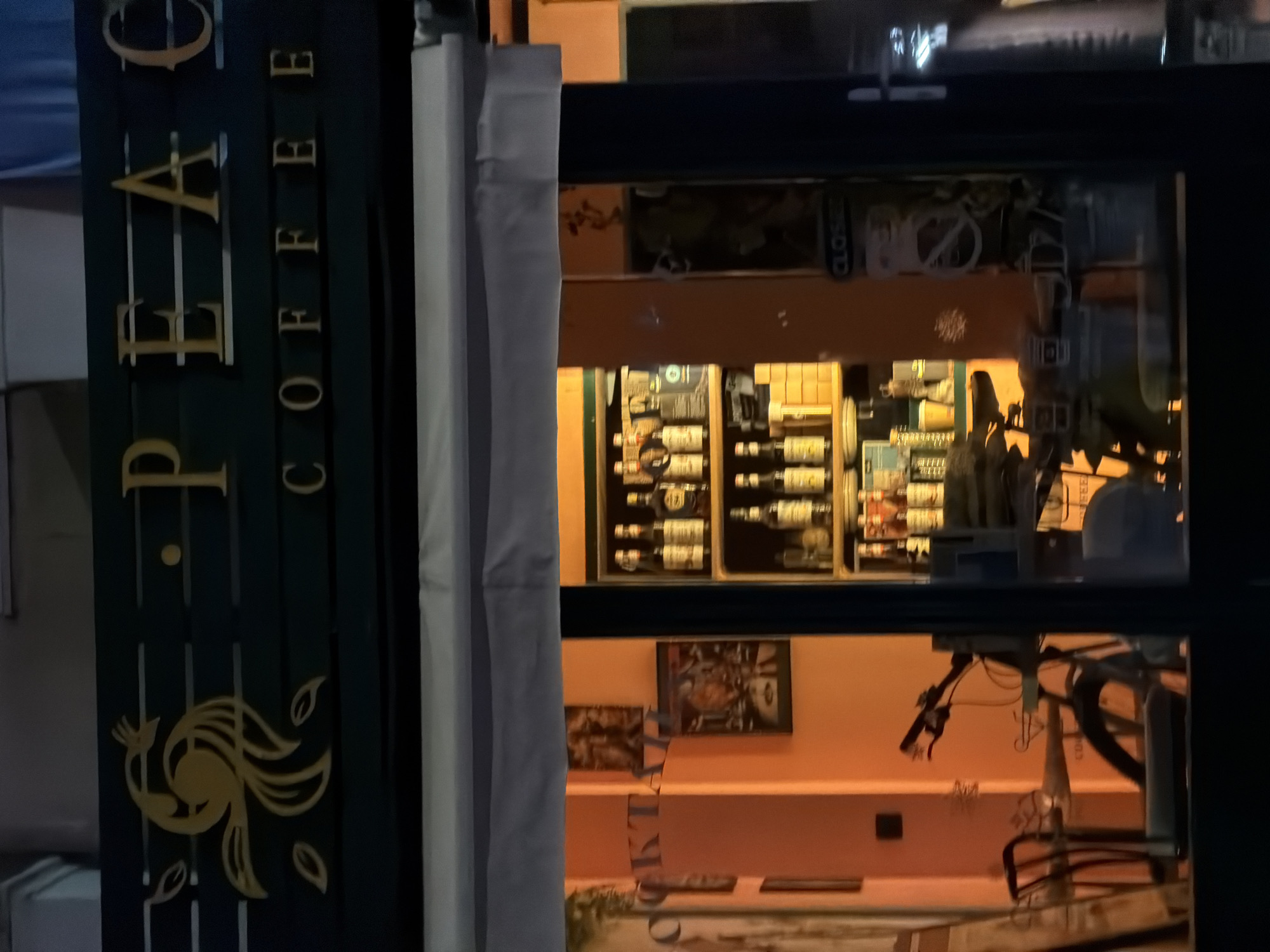 The image shows the exterior of a coffee shop at night. The shop's sign, illuminated in gold lettering against a dark green background, reads "PEACE COFFEE." A stylized gold peacock logo is visible on the left side of the sign. Through the shop's window, the interior is brightly lit, revealing shelves stocked with bottles and other merchandise. A bicycle is parked inside, near a table with chairs. A sign on the door reads "CLOSED." The scene appears to be in Budva, Montenegro.