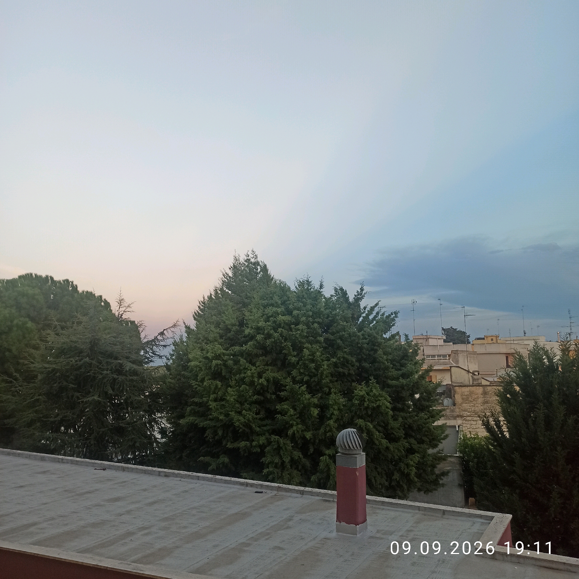The image captures a rooftop view at dusk in Bitonto, Italy. A flat, weathered rooftop occupies the foreground, with a reddish chimney stack featuring a wind-driven turbine vent visible on its surface. Beyond the rooftop, a dense cluster of evergreen trees dominates the mid-ground, their dark foliage contrasting with the sky. In the background, several buildings with light-colored facades and visible antennas are scattered amongst more greenery. The sky transitions from a soft, pale yellow-pink near the horizon to a pale blue, with faint rays of light emanating from an unseen source on the right. A timestamp in the bottom right corner reads "09.09.2026 19:11". There are no people or animals present in the scene.