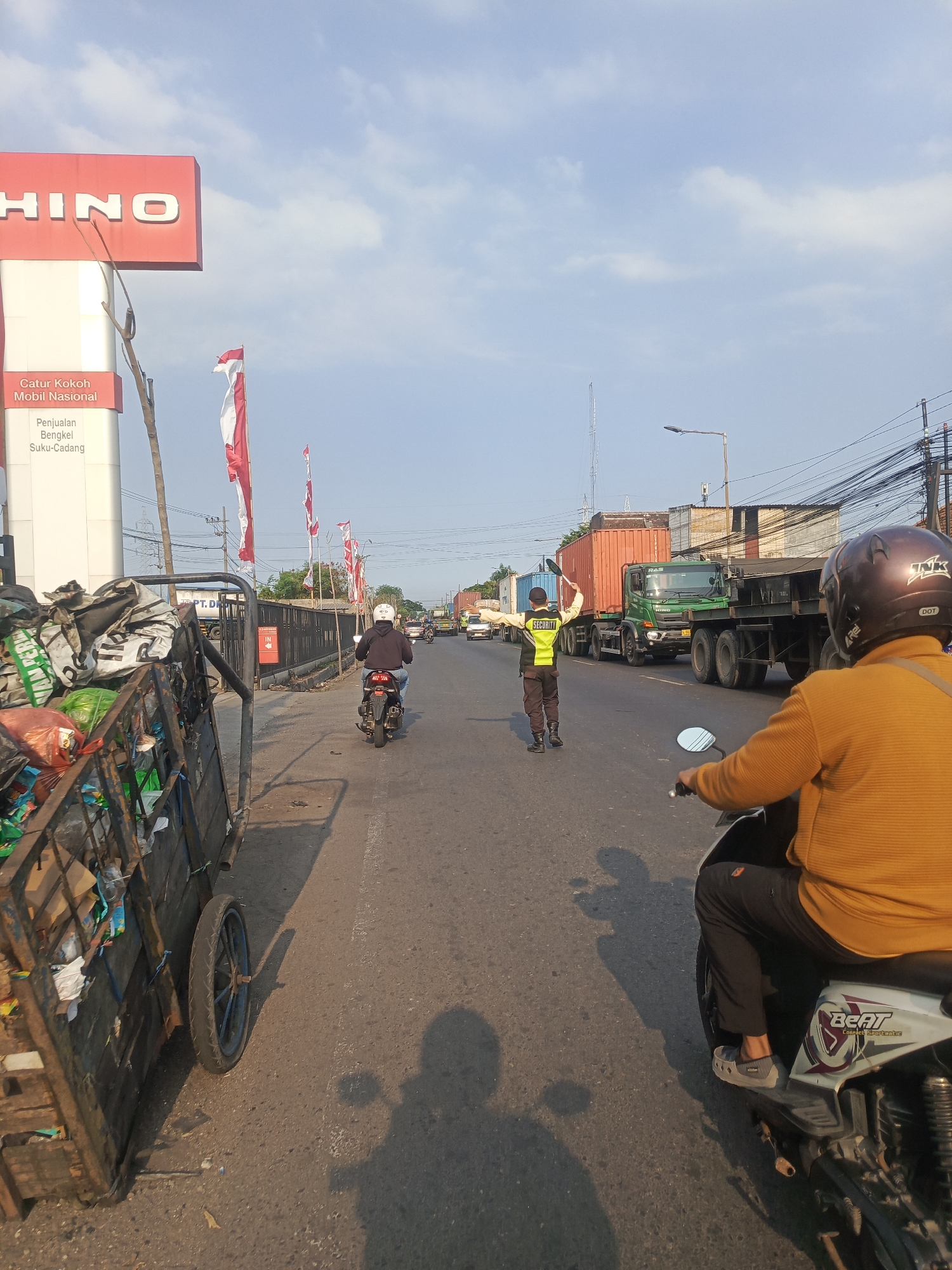This is a vertical, eye-level, medium shot taken from a motorcycle on a street in Surabaya, Indonesia. The foreground is dominated by the rear portion of the motorcycle the photographer is on, with the rider's yellow sweater and helmet visible. To the left, a large, dilapidated wooden cart filled with trash is prominent. The street stretches into the mid-ground and background, with traffic including a motorcycle with a rider in a black hoodie and blue jeans, a security guard in a reflective vest directing traffic, and several large trucks, including a green Hino truck. The sky is a pale blue with scattered clouds. The context of Surabaya, Indonesia is inferred from the visible elements like the Hino dealership sign, Indonesian flags, and common street scenes in that region.