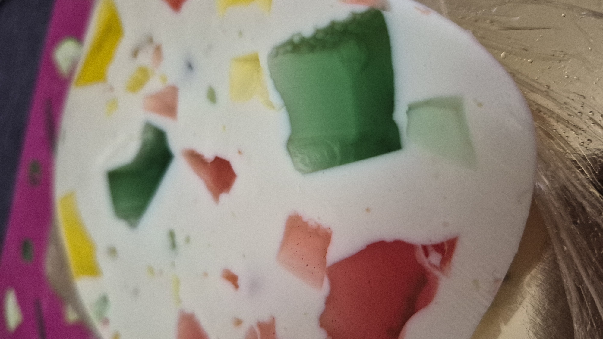 The image shows a close-up view of a dessert, likely a layered gelatin mold, with cubes of red, green, and yellow gelatin suspended in a creamy white base. The dessert is round and appears to be on a golden-brown surface, possibly a plate or tray. In the background, a blurred pink and dark purple patterned surface is visible, which could be a tablecloth or placemat. The lighting is soft, highlighting the textures and colors of the dessert. There is no indication of the city of Krauja, Latvia, in the image.