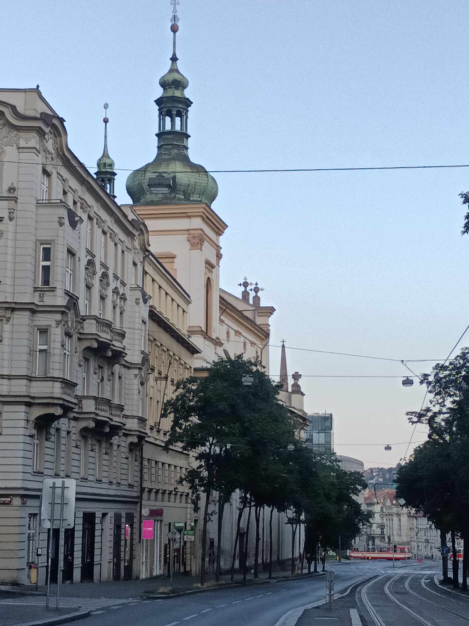 This image captures a street scene in Prague, Czechia. On the left, a row of ornate, historic buildings lines the street, featuring detailed balconies and window frames. Adjacent to these buildings, a church or cathedral with a distinctive green dome and spire rises into the clear sky. The street itself is paved with asphalt and has tram tracks running through it. In the distance, more buildings are visible, along with a red tram. Several trees with dark green foliage are interspersed along the street, casting some shadows. The sky is a pale blue, indicating it is likely early morning or late afternoon. Overhead wires, likely for the tram system, crisscross the scene.