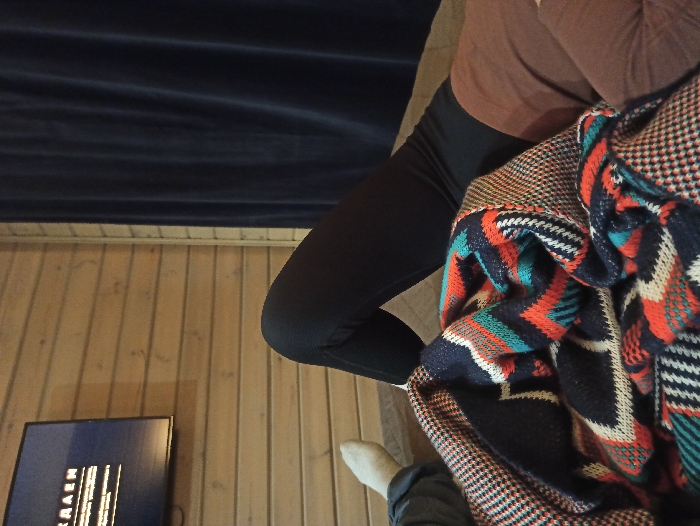 An indoor scene in Nizhneye Zadolgoye, Russia, depicts at least one individual resting. A person is observed lying on a light-brown surface, partially covered by a patterned blanket. This blanket features navy blue, orange, teal, and white geometric designs, with a brown and beige houndstooth pattern visible on its edge. The individual wears black leggings on their bent left leg and a brown top. A second person's left foot, clad in a white sock, is partially visible on the far left. In the upper-left quadrant, a flat-screen television is mounted on a wall constructed of horizontal light-toned wooden planks, displaying white text, including partial Russian characters "КЛАДЕН", against a dark background. A dark blue curtain hangs vertically on the right side of the frame.