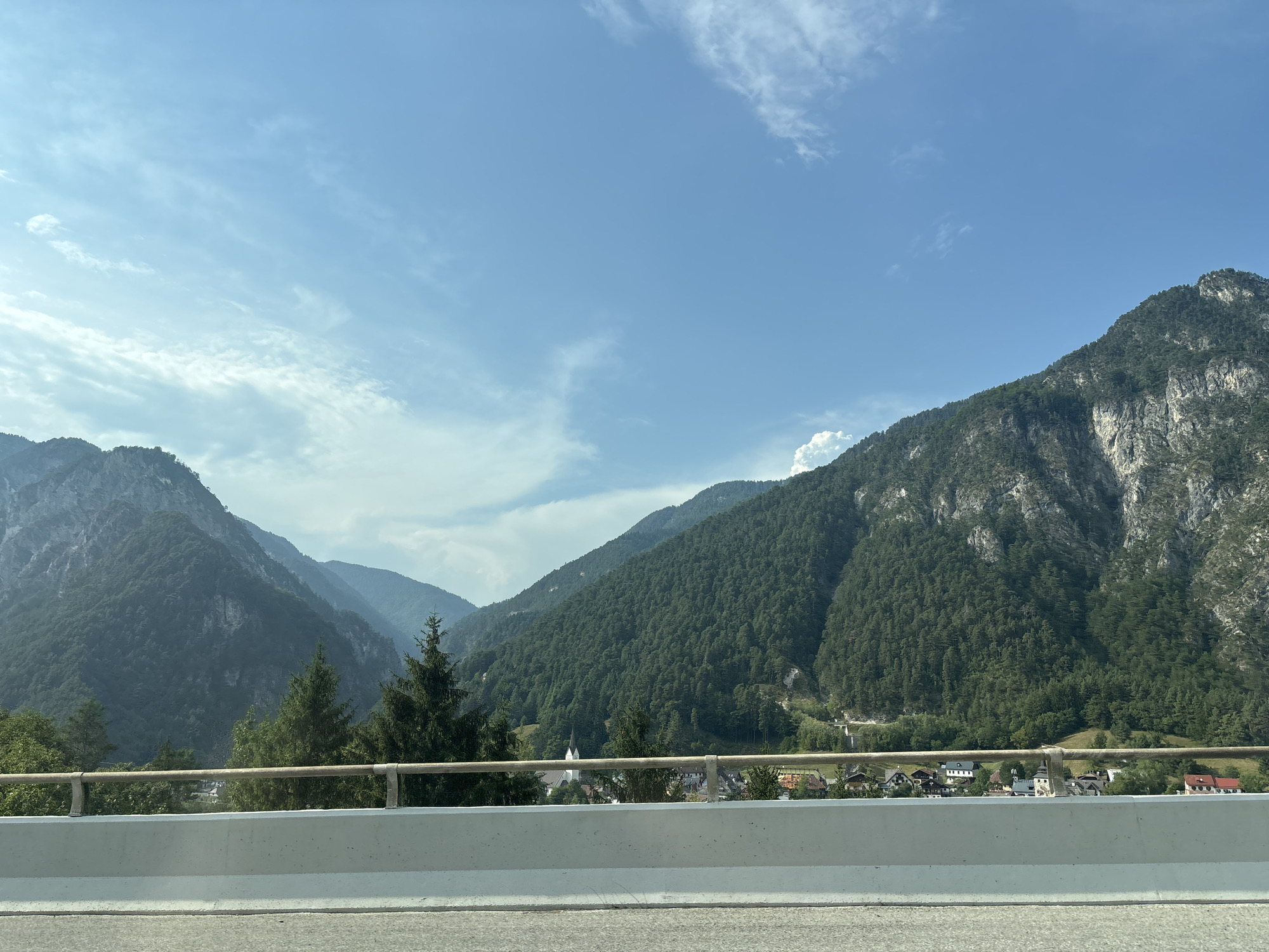 The image shows a scenic view of a valley in Malborghetto-Valbruna, Italy, with a clear blue sky and scattered clouds. In the foreground, there is a concrete barrier and a metal railing, suggesting the photo was taken from a highway or elevated viewpoint. Beyond the barrier, a small village is nestled in the valley, characterized by traditional houses with red and white roofs, and a prominent white church steeple. The village is surrounded by steep, densely forested mountains. The trees on the mountainside are predominantly evergreen, creating a lush green appearance. The scale of the mountains is impressive, with rugged rocky outcrops visible on the right side of the frame. The lighting suggests it is daytime, likely late morning or early afternoon, with the sun casting shadows and highlighting the textures of the mountains and foliage. The overall impression is one of natural beauty and a peaceful, rural setting.