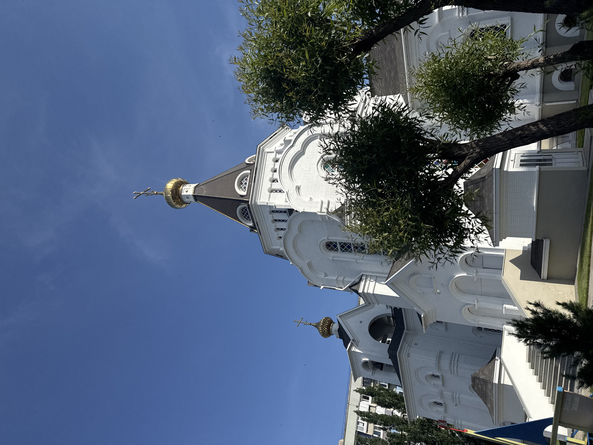 A low-angle, medium shot depicts a white Orthodox church against a blue sky. The church is adorned with golden domes and crosses, and a few trees frame the structure. The location appears to be urban, with the church situated near a building, possibly in the city of Unknown, Unknown. No people are visible in the frame.