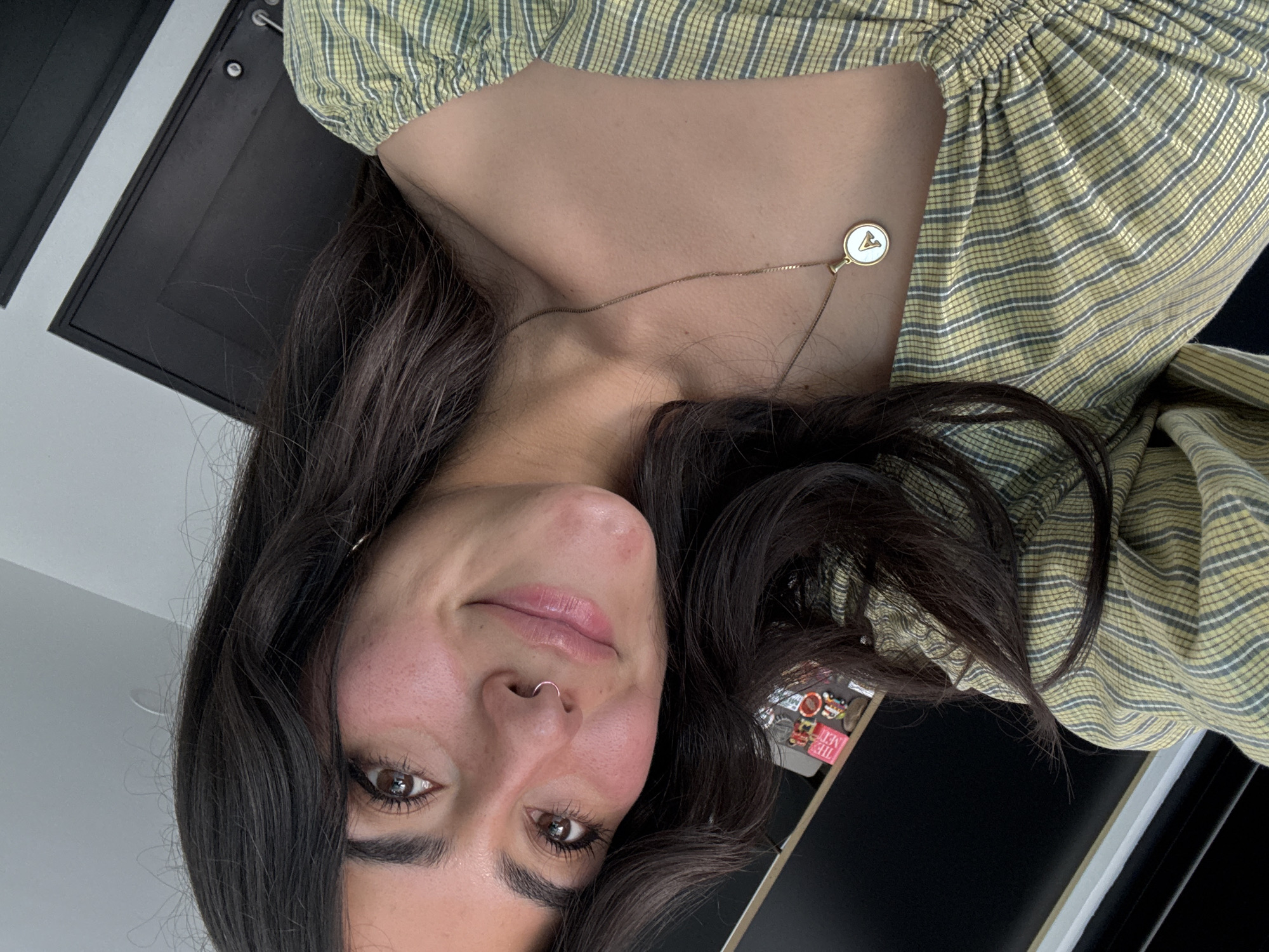A young woman with dark hair, wearing a yellow and grey striped shirt and a necklace with a round pendant, is shown in a close-up shot. She has brown eyes, a nose ring, and is smiling slightly. The background includes a black surface, potentially a desk, with small objects visible on it. The location is not discernible from the image; therefore, the city of San Bernardino Tlaxcalancingo, Mexico, cannot be confirmed.