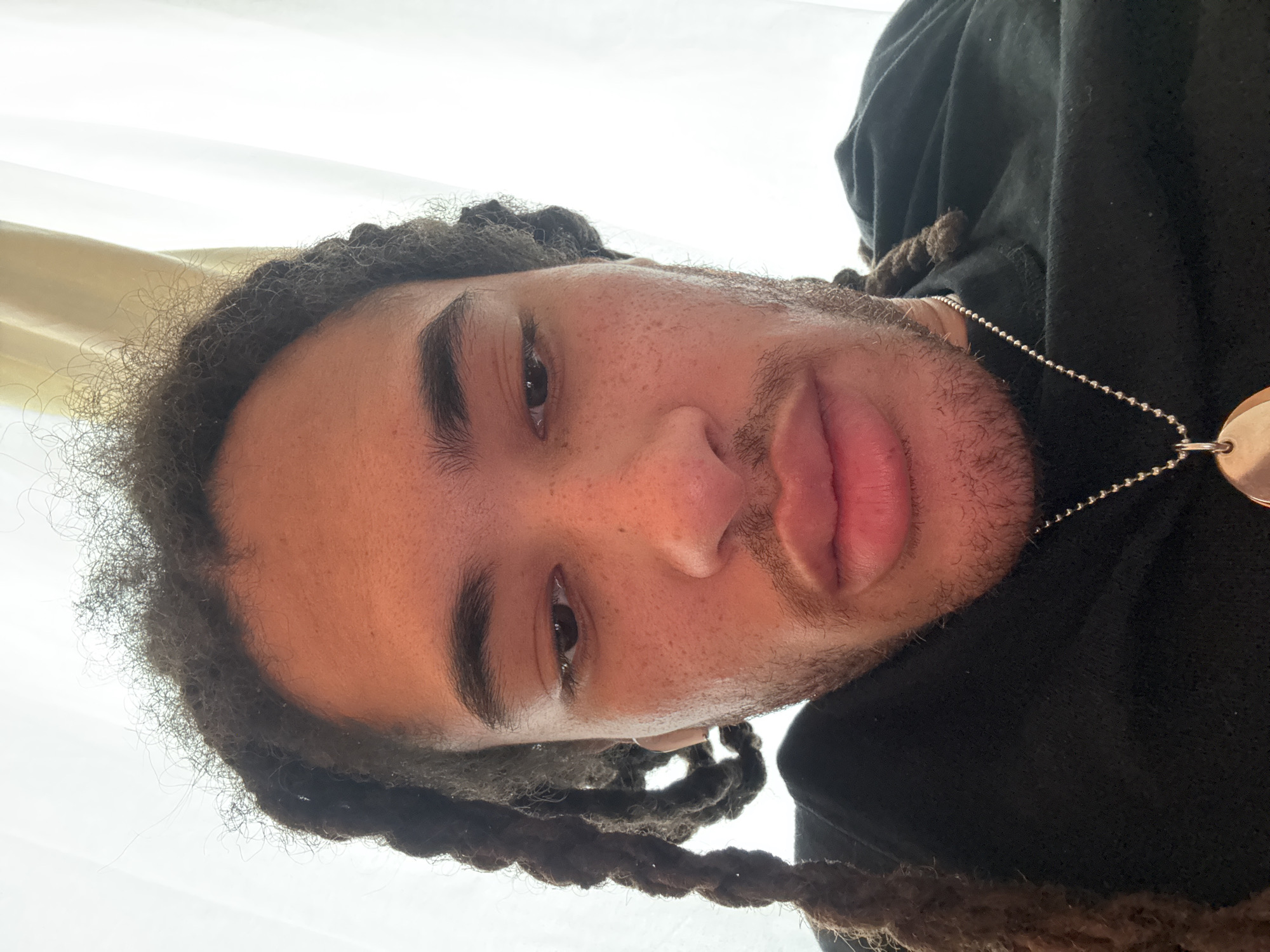 A close-up, angled selfie of a person with dark skin, dreadlocks, and a chain necklace, wearing a black hoodie. The person is looking down at the camera with a neutral expression. The background consists of a beige wall and a window with white curtains. The location is not discernible from the image.