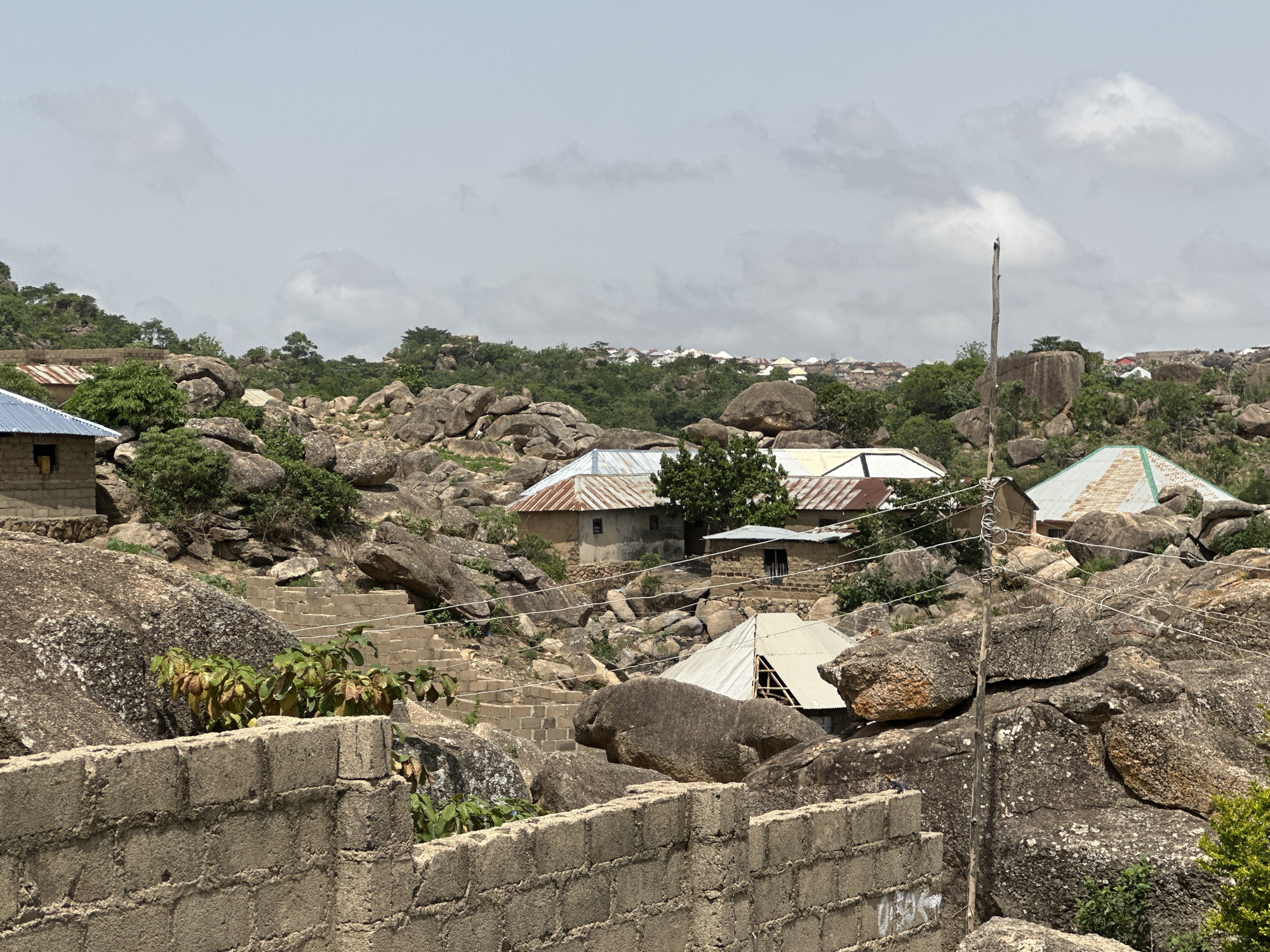 The image depicts a residential area in Jos, Nigeria. The landscape is rocky, with numerous large boulders interspersed among the houses. Several single-story dwellings are visible, with corrugated metal roofs in various states of repair. Some houses appear to be under construction. The houses are constructed from concrete blocks. A retaining wall made of concrete blocks runs along the lower foreground. Electrical wiring is strung between poles and buildings. The sky is overcast. Vegetation is sparse, consisting primarily of small trees and shrubs.