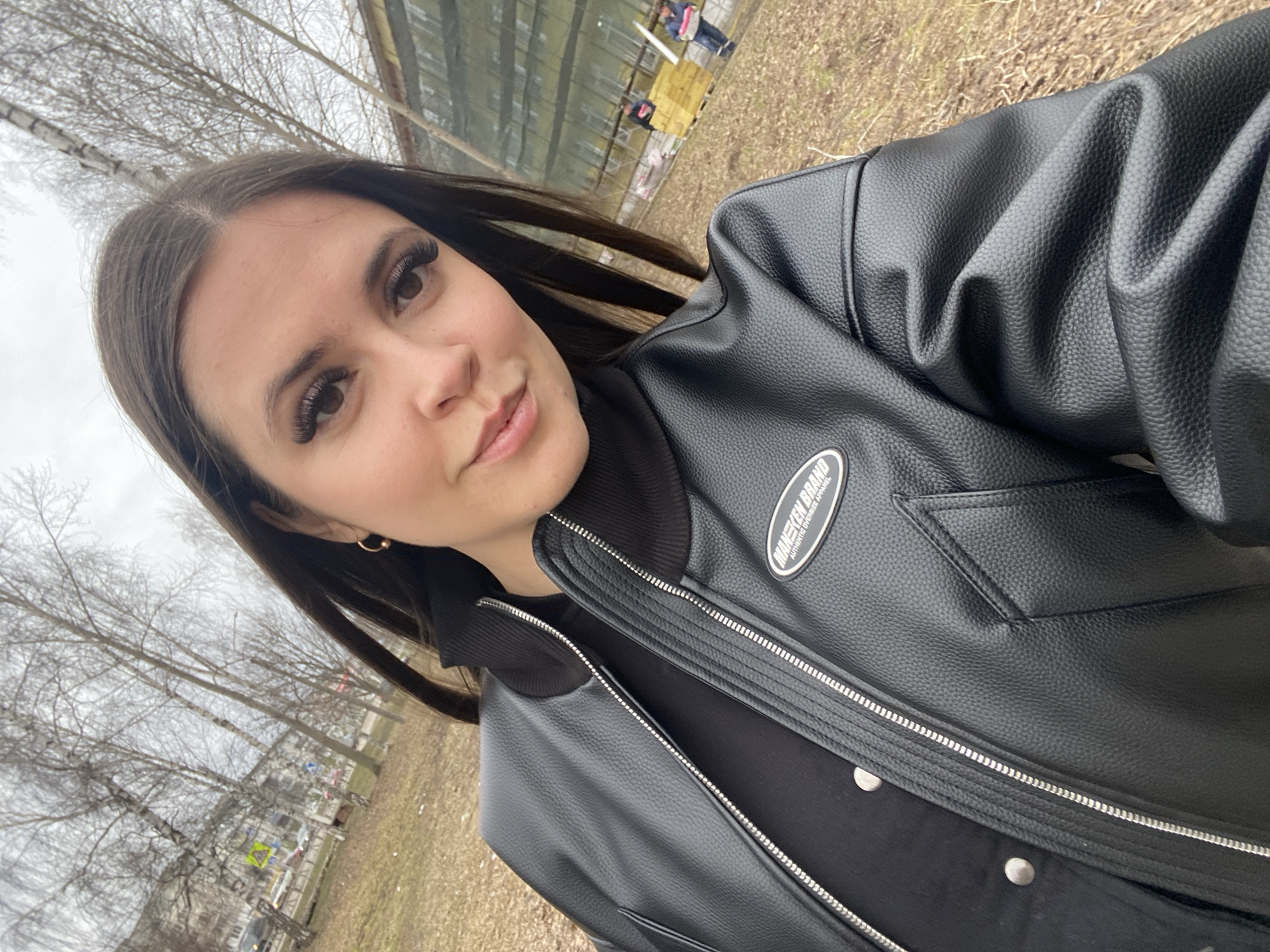 A young woman is taking a selfie outdoors in what appears to be a park or green space. She has long, dark hair, dark eyes, and is wearing artificial eyelashes. Her skin is fair. She is wearing a black leather-look bomber jacket over a black turtleneck sweater. The jacket has a silver zipper. The photo is taken at an angle, and the background shows a row of bare trees, brown grass, and a building in the distance. Based on the presence of birch trees, the location is likely Russia, specifically the city of Chaikovsky. There is evidence of people in the background as well as street signs.