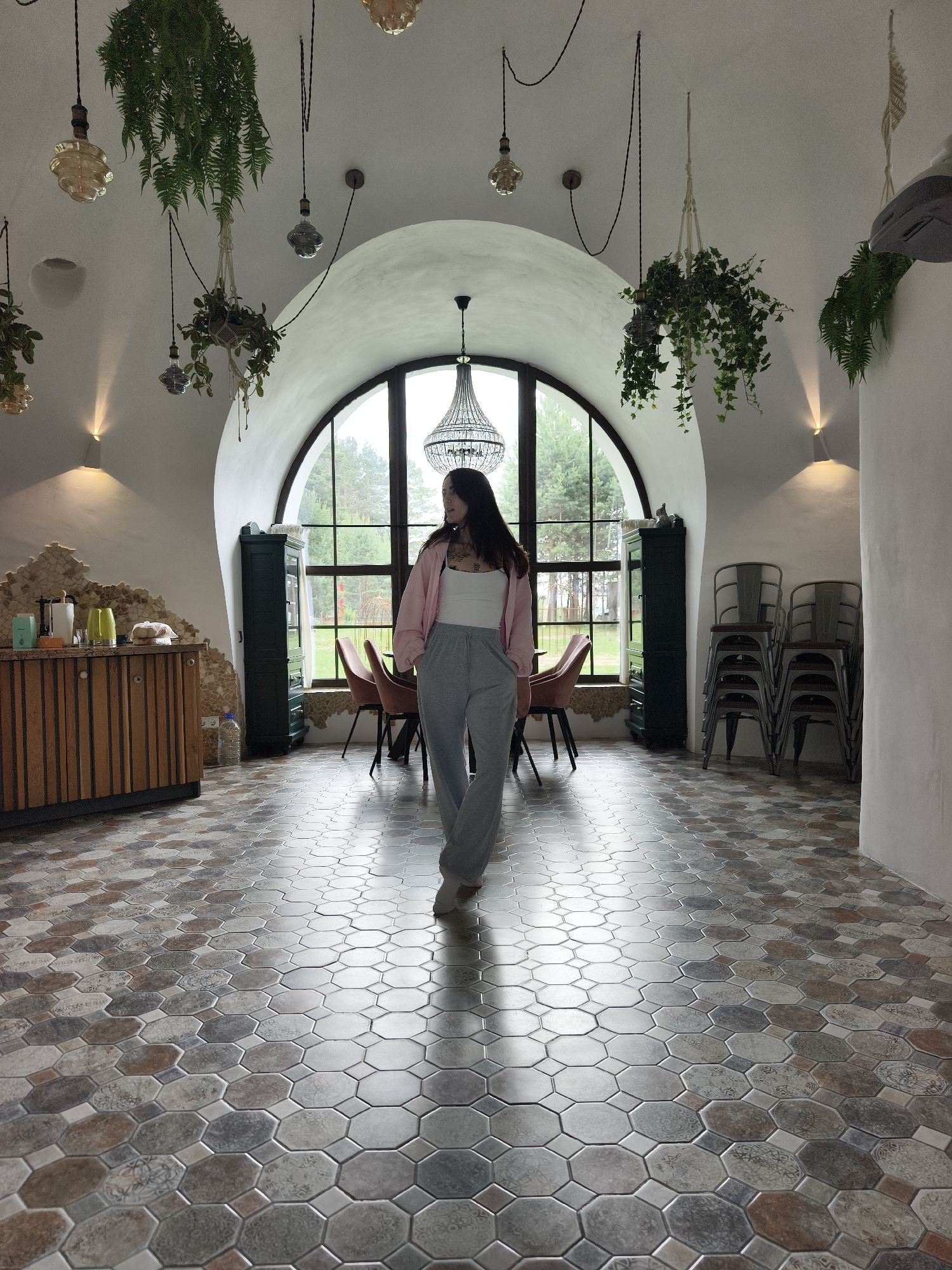 A young woman with dark hair, wearing a white tank top, a pink cardigan, and grey sweatpants, walks across a room with an octagonal tiled floor. She is positioned in the center of the frame, looking to her left. The room features a large arched window with a view of trees outside, suggesting a natural setting. Hanging plants are suspended from the ceiling, adding a touch of greenery to the interior. Several decorative light fixtures are also visible.
To the left of the woman, there is a wooden cabinet with stone accents and various items on top, including what appear to be bottles and containers. Further left, a tall, dark green cabinet stands near the window. To the right of the woman, stacks of metal chairs are visible. Pink upholstered chairs are placed near the window.
The lighting in the room is soft, with wall sconces casting light upwards. The scene appears to be indoors, possibly a cafe, restaurant, or a unique living space. The overall ambiance is relaxed and casual, with the woman's attire and pose suggesting a moment of leisure. There is no visible text in the image. The time of day appears to be daytime, and the weather outside seems overcast or mild, based on the natural light coming through the window.