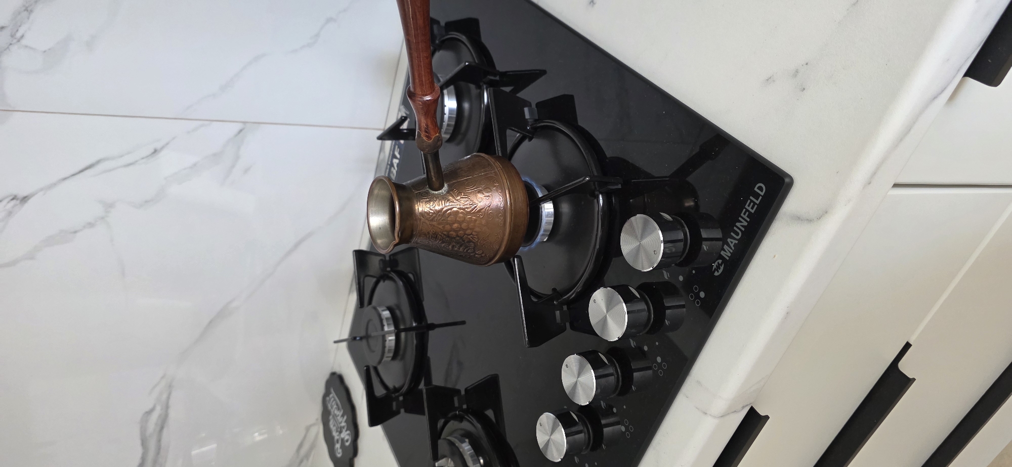 An overhead medium shot captures a copper cezve being heated on a black gas stove in a kitchen. The cezve, adorned with intricate grape leaf engravings, sits on a burner with a visible blue flame. Surrounding the cezve are four chrome knobs on the glass-top stove, each with a circular pattern. The stove itself is embedded in a white marble countertop. Behind the stove, a white tiled backsplash with subtle grey veining extends upwards. To the left of the stove, a small, dark chalkboard sign with white lettering is partially visible. The overall setting suggests a domestic kitchen environment.