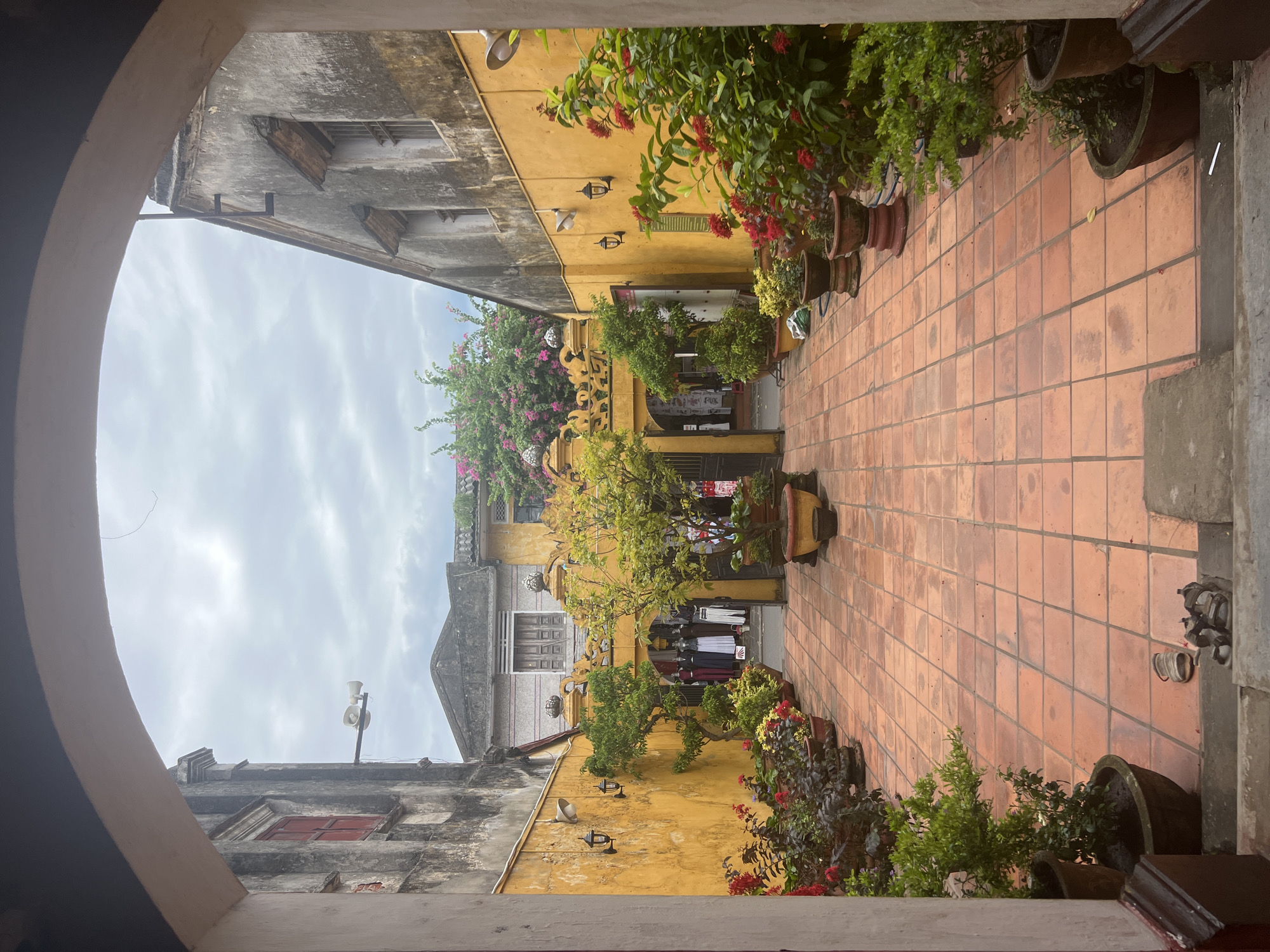 This is a photograph taken from a covered archway looking into a courtyard in Hoi An, Vietnam. The courtyard is paved with terracotta tiles and is lined with buildings on either side. The buildings are old, with weathered yellow and grey walls. There are potted plants and flowers around the courtyard, and an archway in the center leads to what appears to be a shop. The sky is overcast. There are no people visible in the photograph.