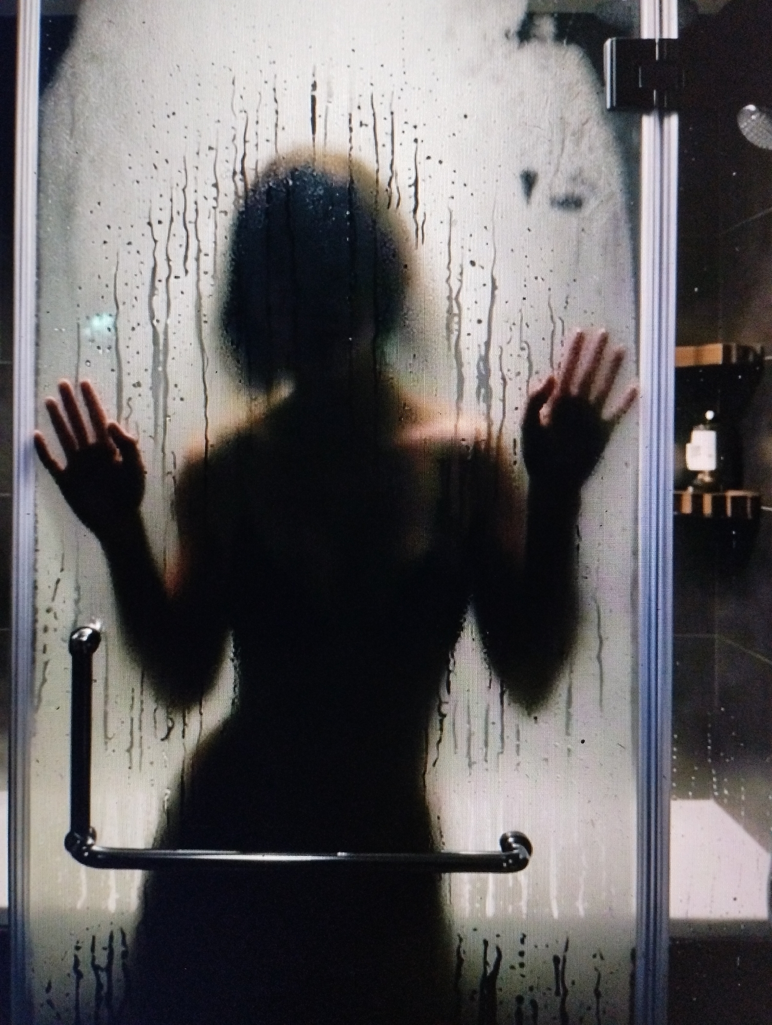 A silhouetted figure, appearing to be an adult, stands behind a steamy glass shower door. Their hands are pressed against the glass, one on the left and one on the right, with fingers splayed. Water droplets and streaks are visible on the glass, obscuring finer details of the figure. The figure's head and shoulders are discernible as a dark mass. A metal shower door handle is visible on the left side. The background is dark and out of focus, with hints of shelves and objects that suggest a bathroom environment.
